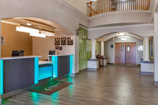 A hotel reception area with a reception desk, a digital display, framed awards on the wall, and a staircase leading to an upper level.