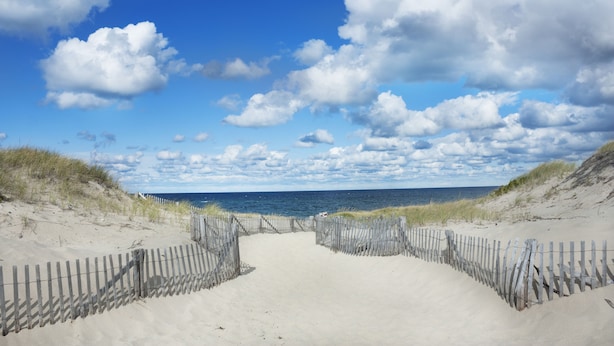 A sandy beach path with a wooden fence leading to the ocean, under a partly cloudy sky.