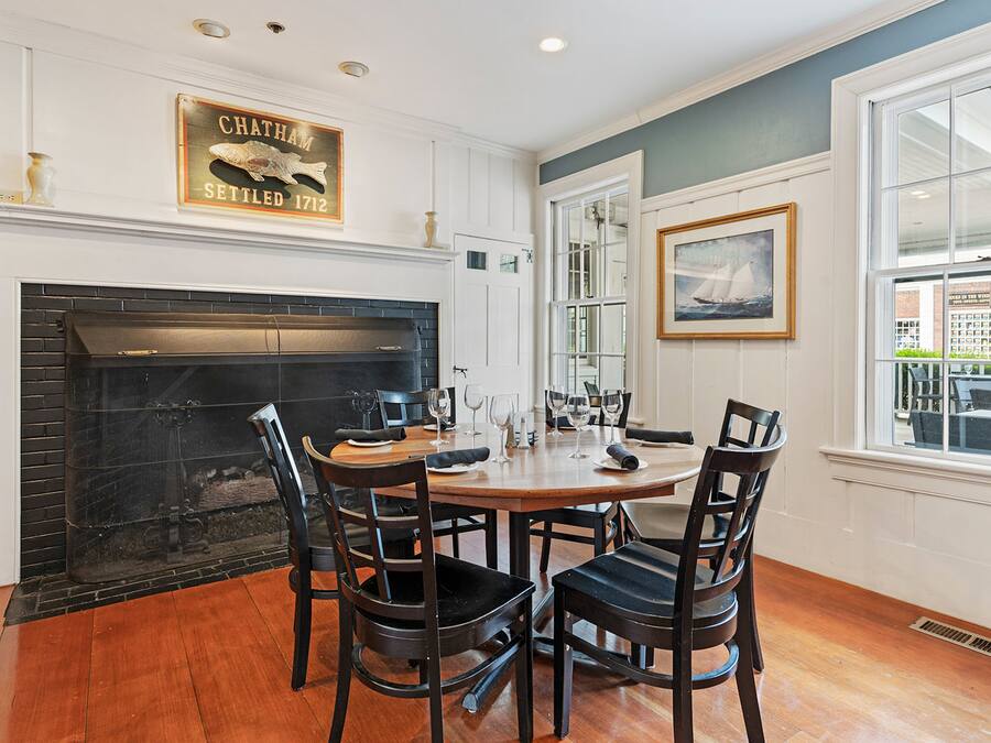 A dining area with a fireplace, wooden chairs, a round table, and a framed picture on the wall.