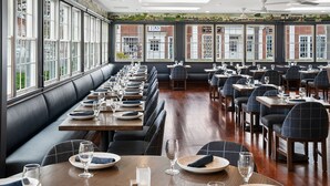 A restaurant with long communal tables and booths, large windows, and a view of buildings outside.