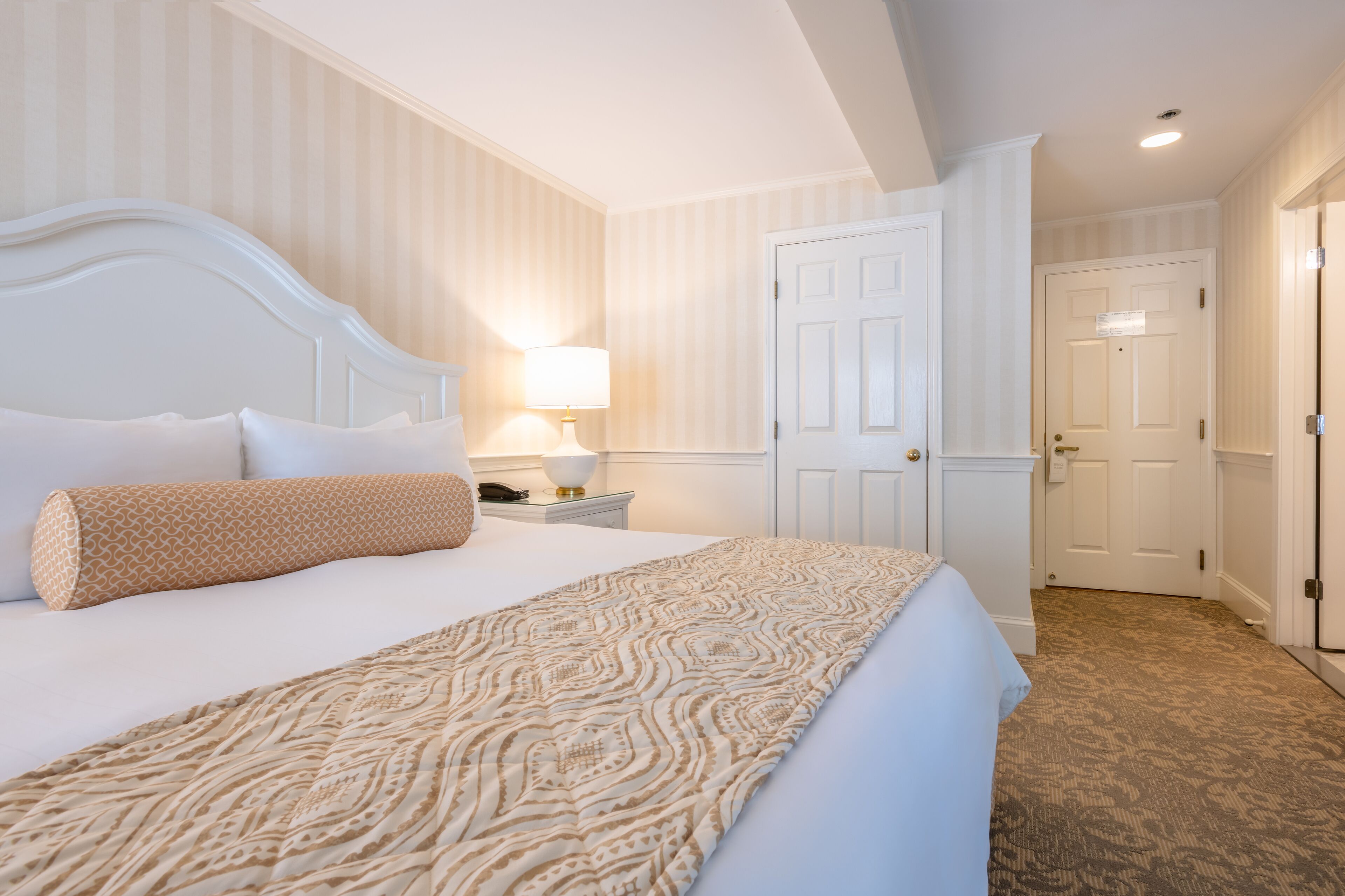 A hotel room with a large bed, a bedside table with a lamp, a door, and a carpeted floor.