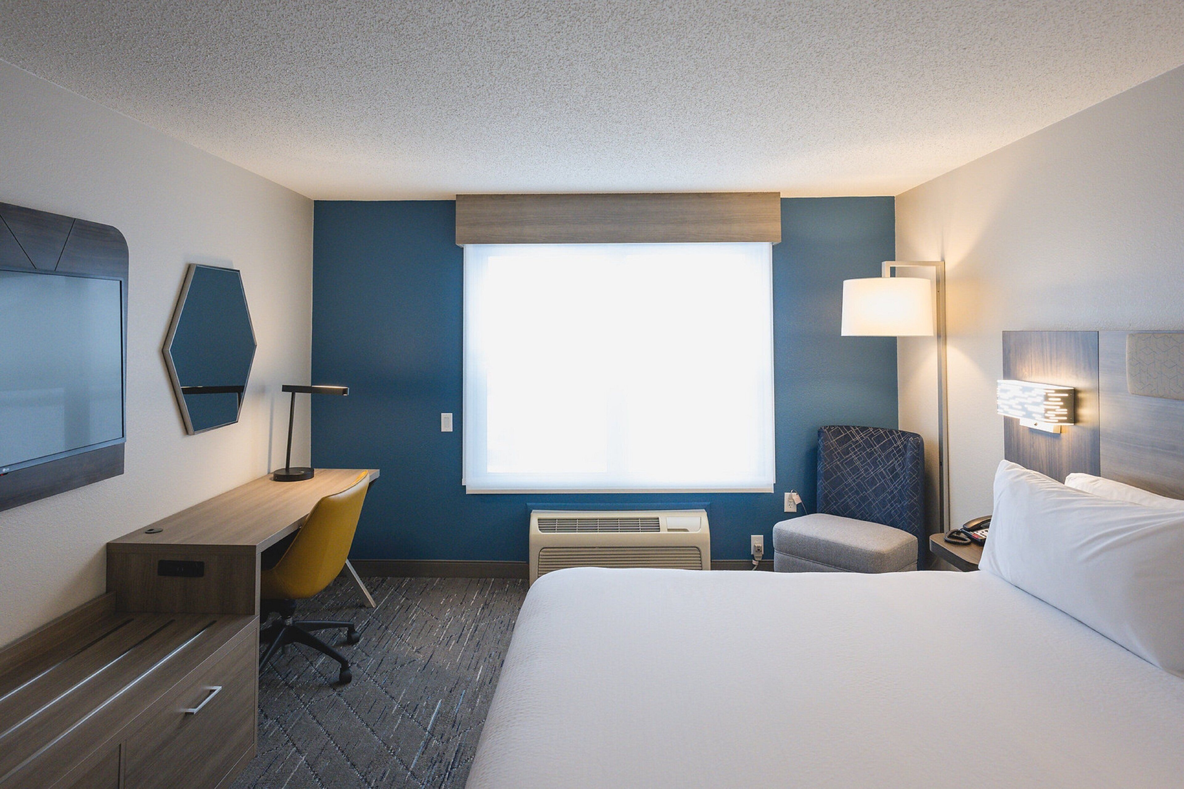 Quarto de hotel com cama, escrivaninha, cadeira, televisão, janela e luminária.
