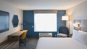 Quarto de hotel com cama, escrivaninha, cadeira, televisão, janela e luminária.