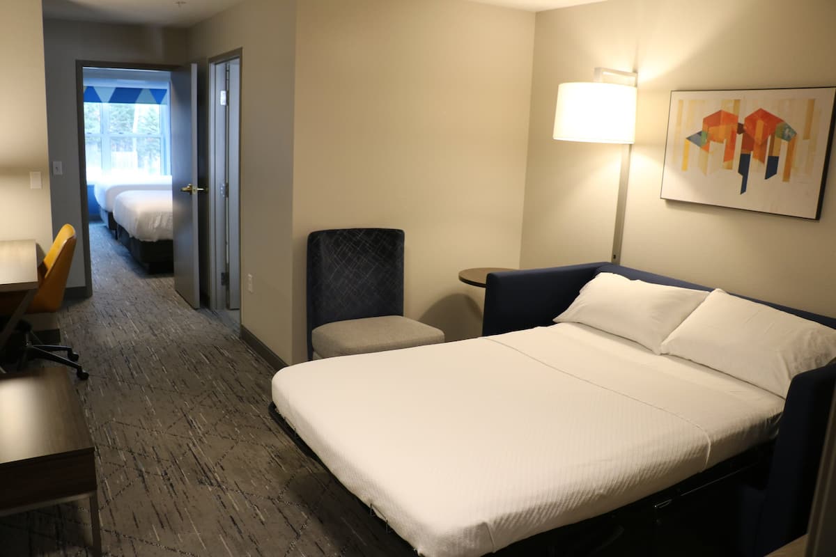 Habitación de hotel con una cama grande, un escritorio, una silla y una lámpara. Se ve una puerta que conduce a otra habitación con una cama.