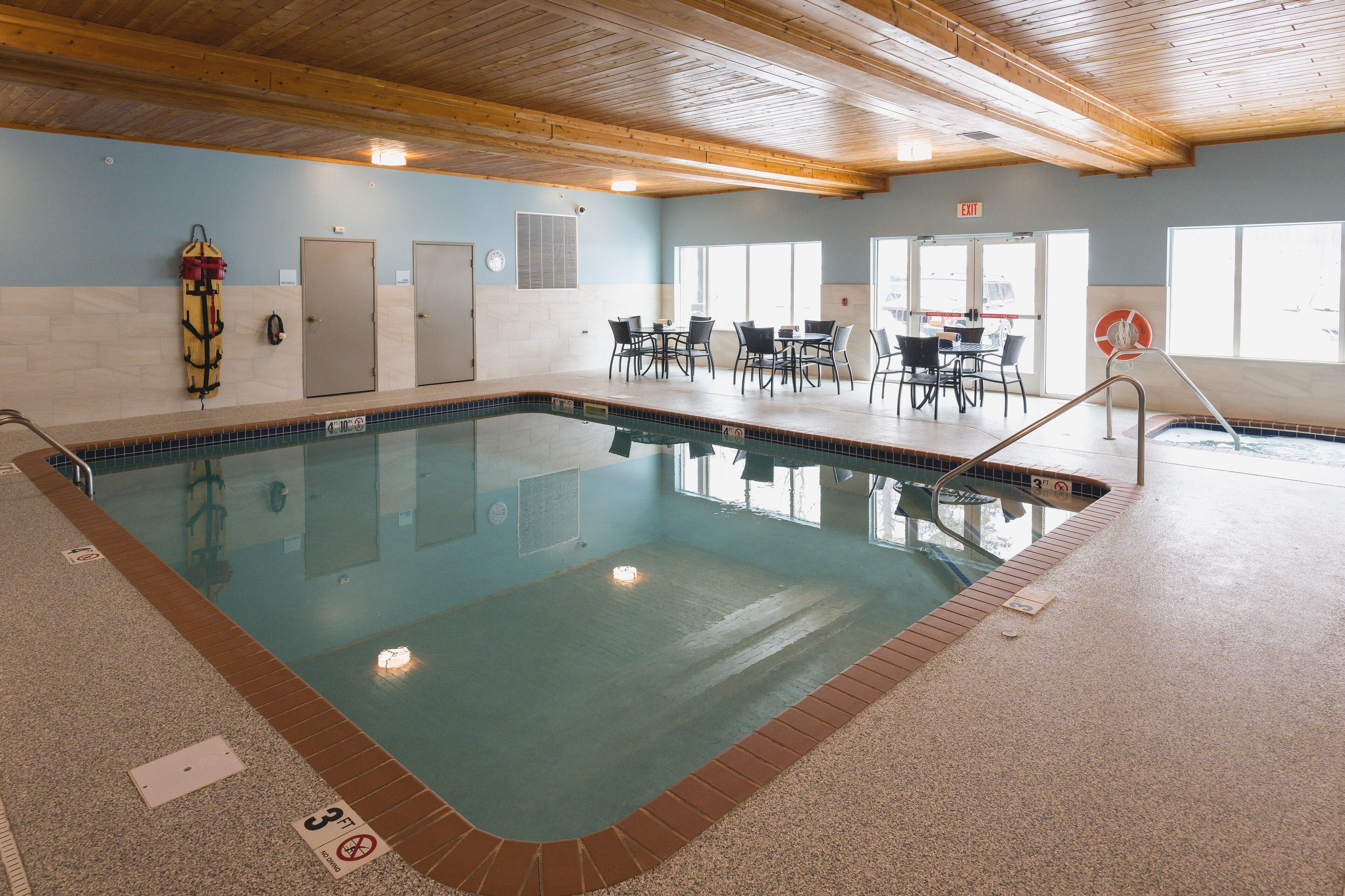 Una piscina interior con borde de azulejos, vigas de madera en el techo y una zona de estar con mesas y sillas.