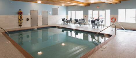 Una piscina interior con borde de azulejos, vigas de madera en el techo y una zona de estar con mesas y sillas.