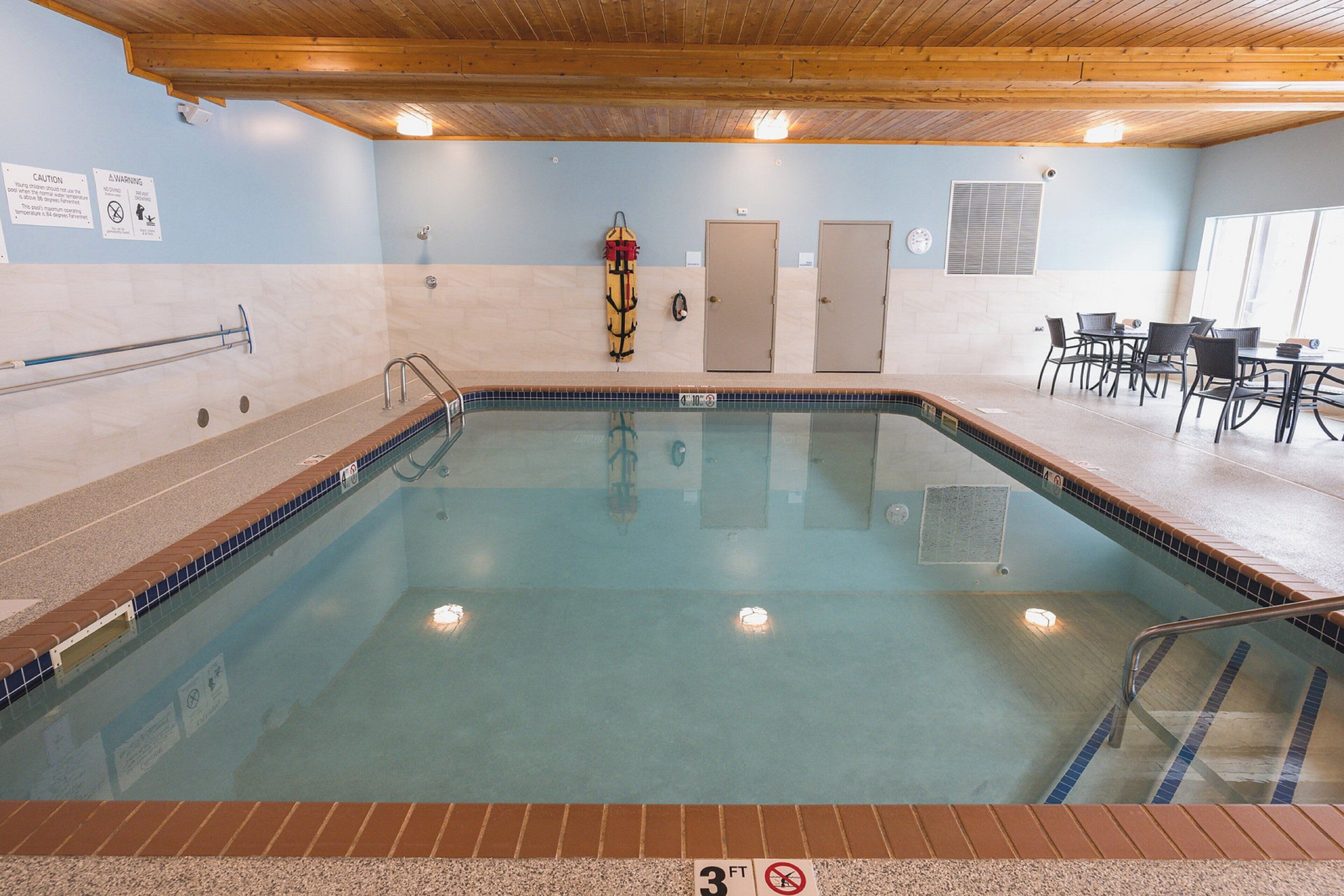 An indoor swimming pool with a 3ft depth sign, a lifeguard stand, and a seating area with tables and chairs.