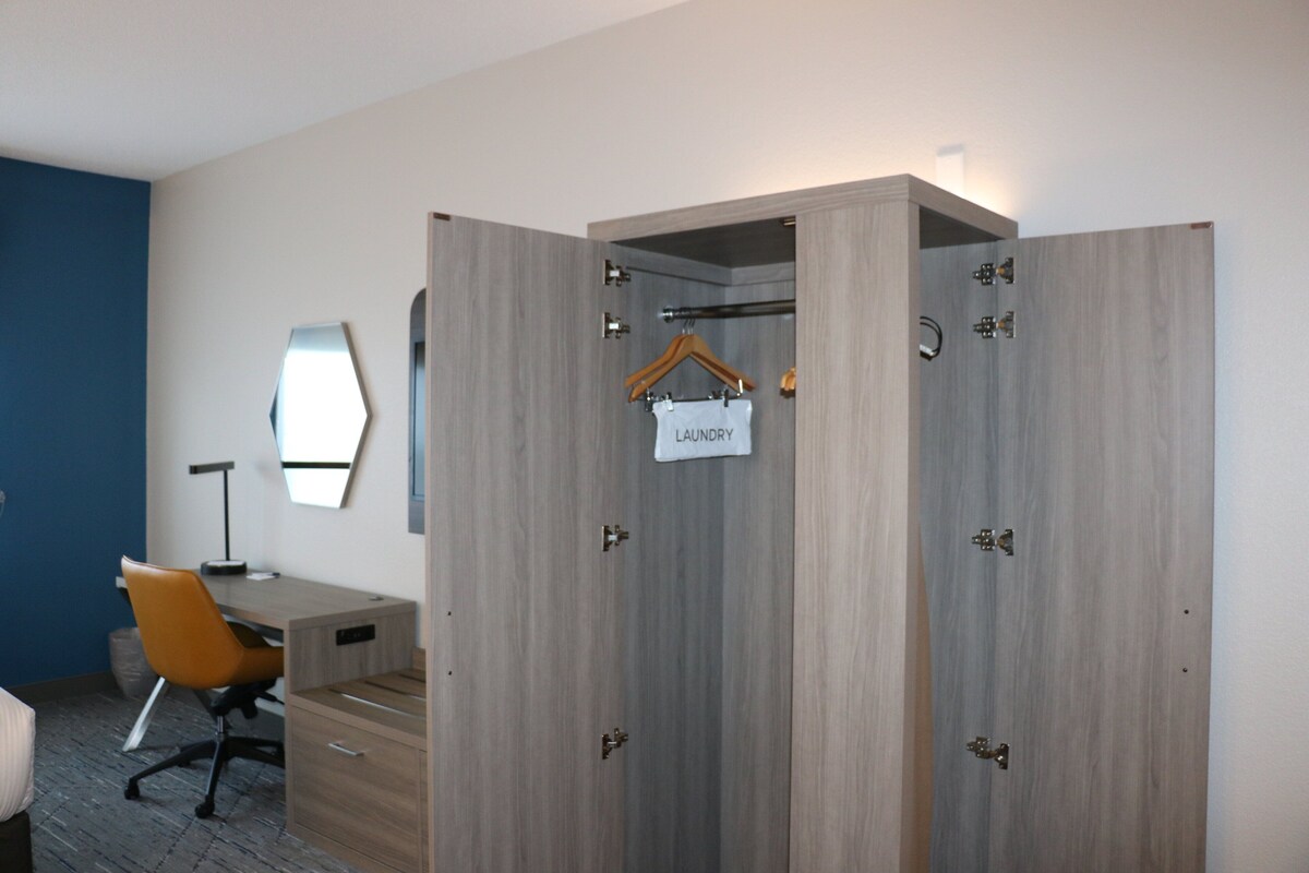 Une chambre d’hôtel avec une armoire en bois, un bureau, une chaise orange et un miroir au mur.