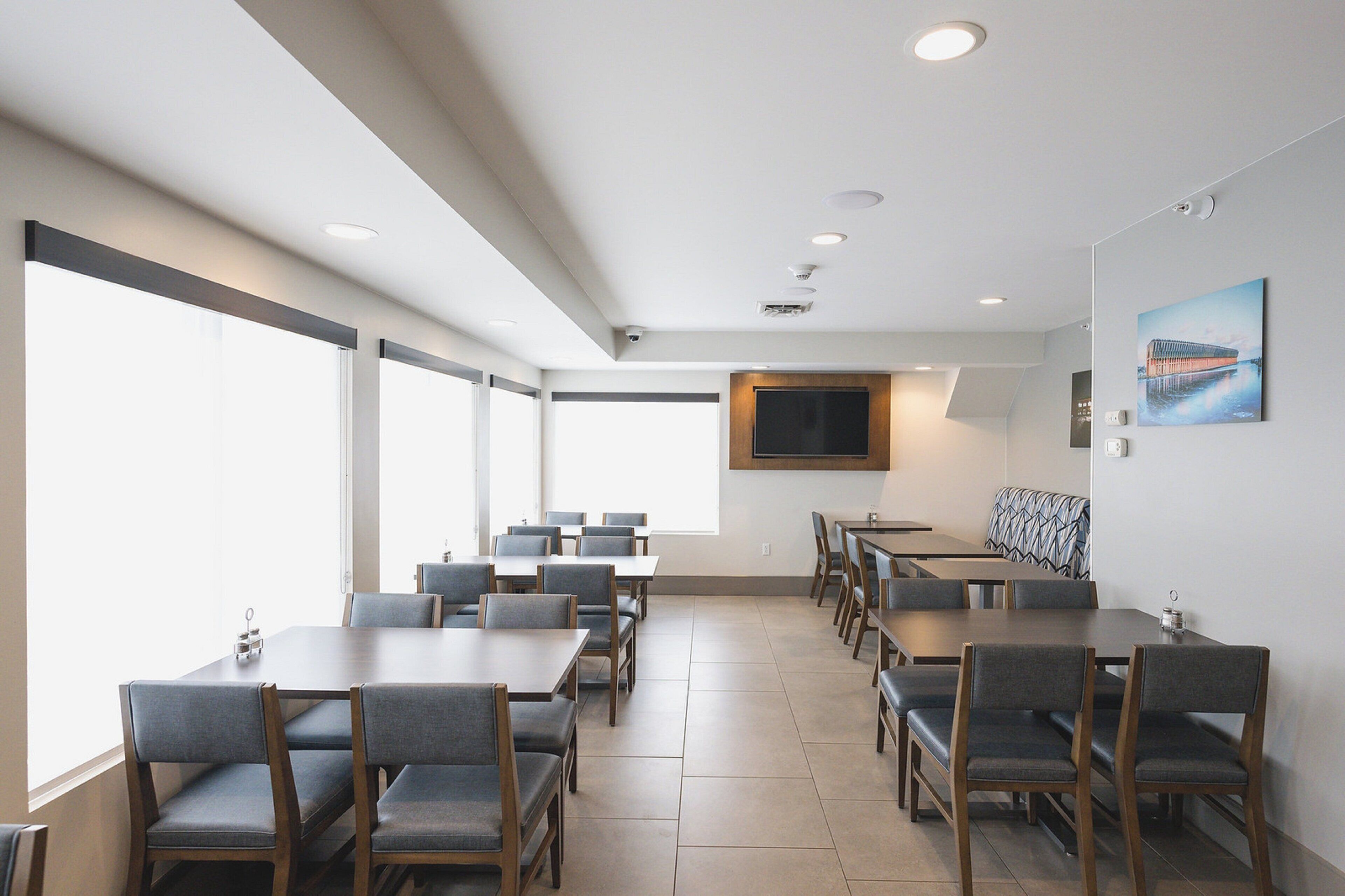 A conference room with multiple tables and chairs, large windows, a mounted television, and a painting on the wall.