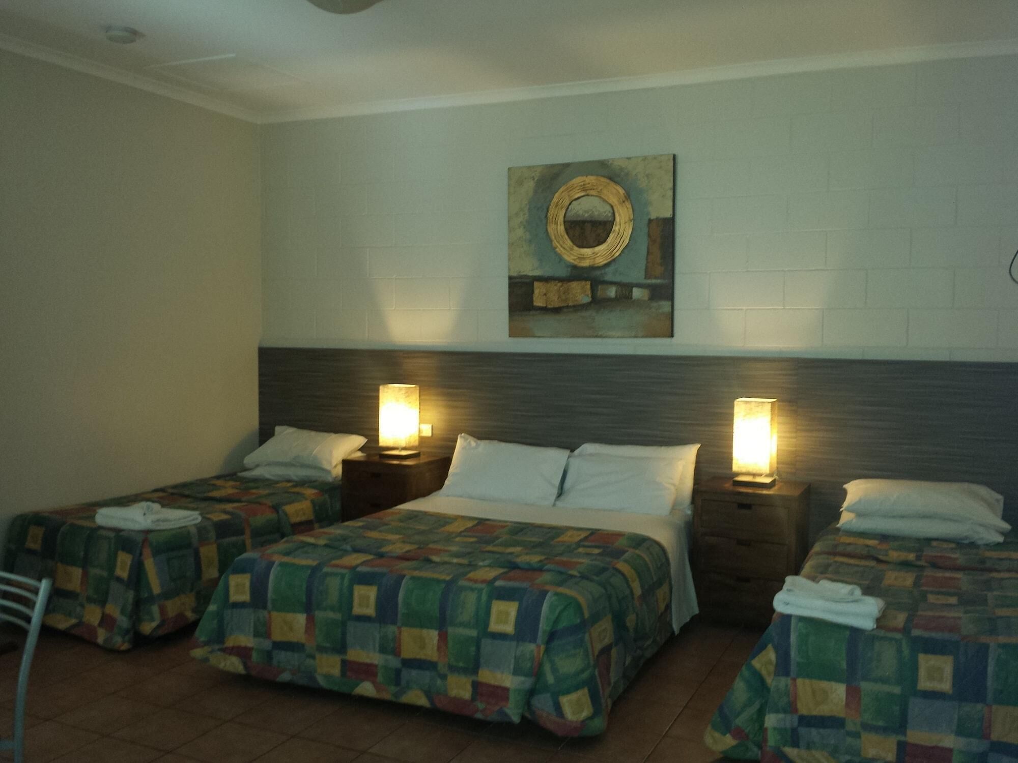 A hotel room with three beds, each with a quilted bedspread and a pillow. There are two bedside lamps and a wall-mounted mirror above the beds.