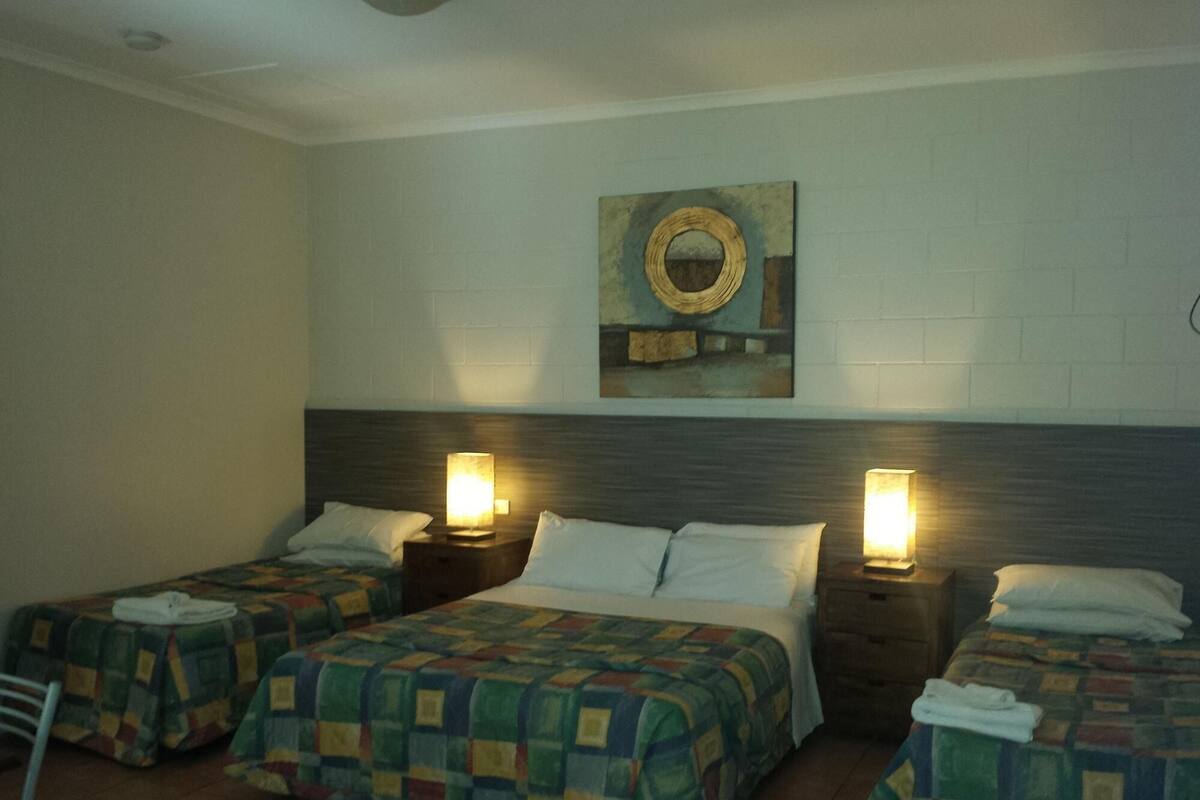 A hotel room with three beds, each with a quilted bedspread and a pillow. There are two bedside lamps and a wall-mounted mirror above the beds.