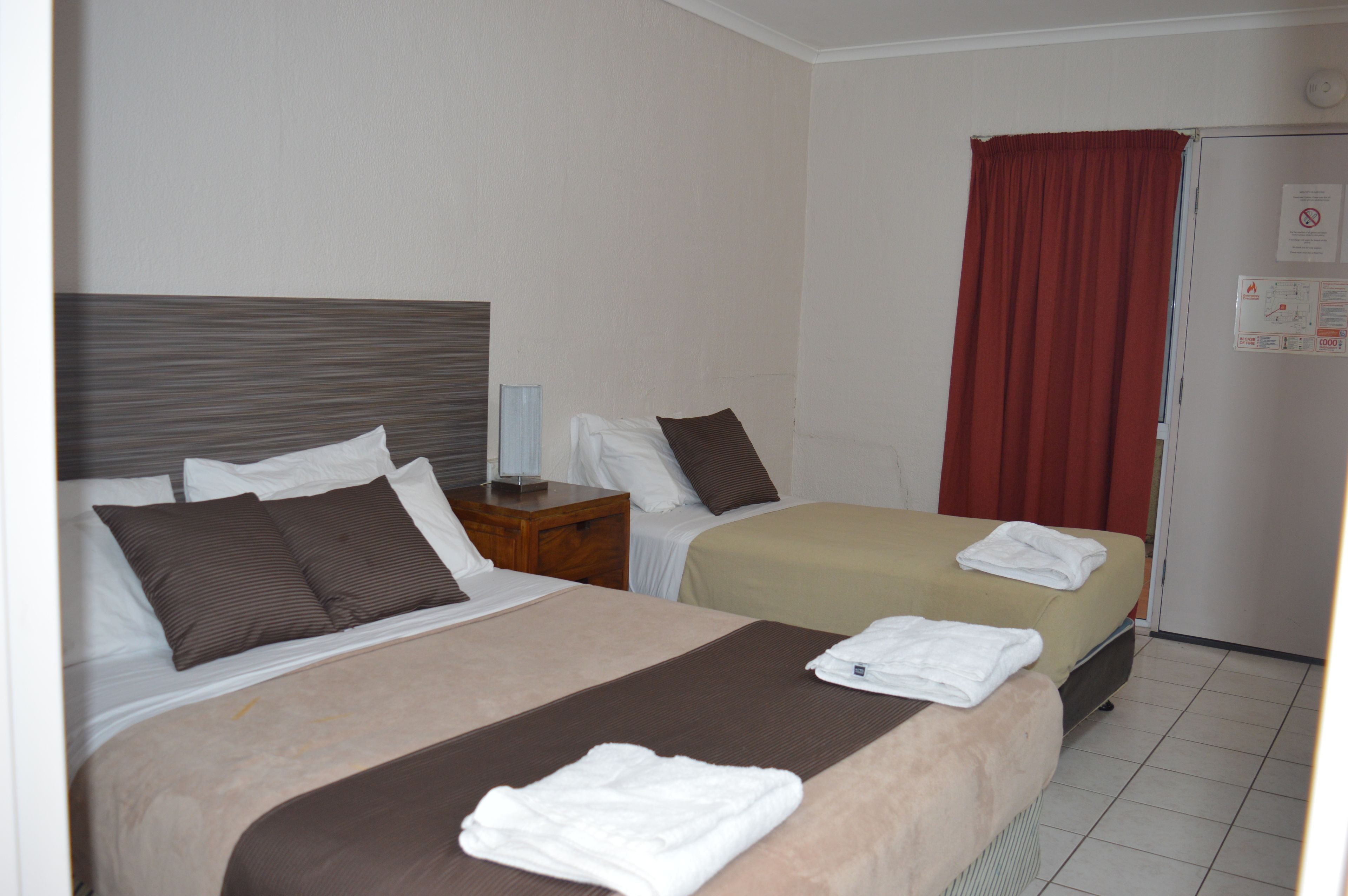 Une chambre d’hôtel avec deux lits, chacun doté d’une tête de lit marron, de draps blancs et d’un rideau rouge. On y trouve une petite table avec une lampe et une horloge au mur.
