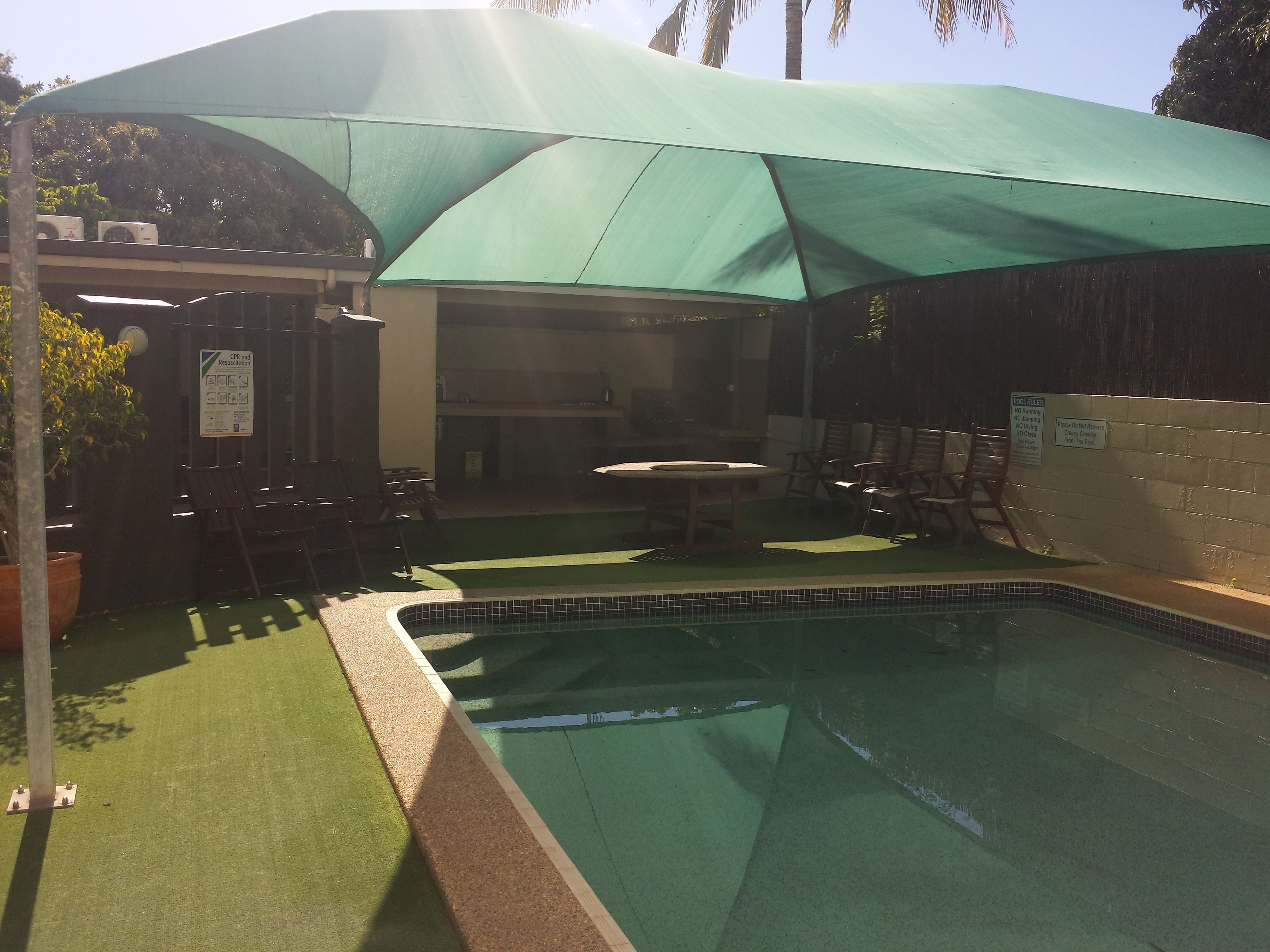 Une piscine avec un auvent vert pour créer de l’ombre, une table et des chaises placées à l’ombre.