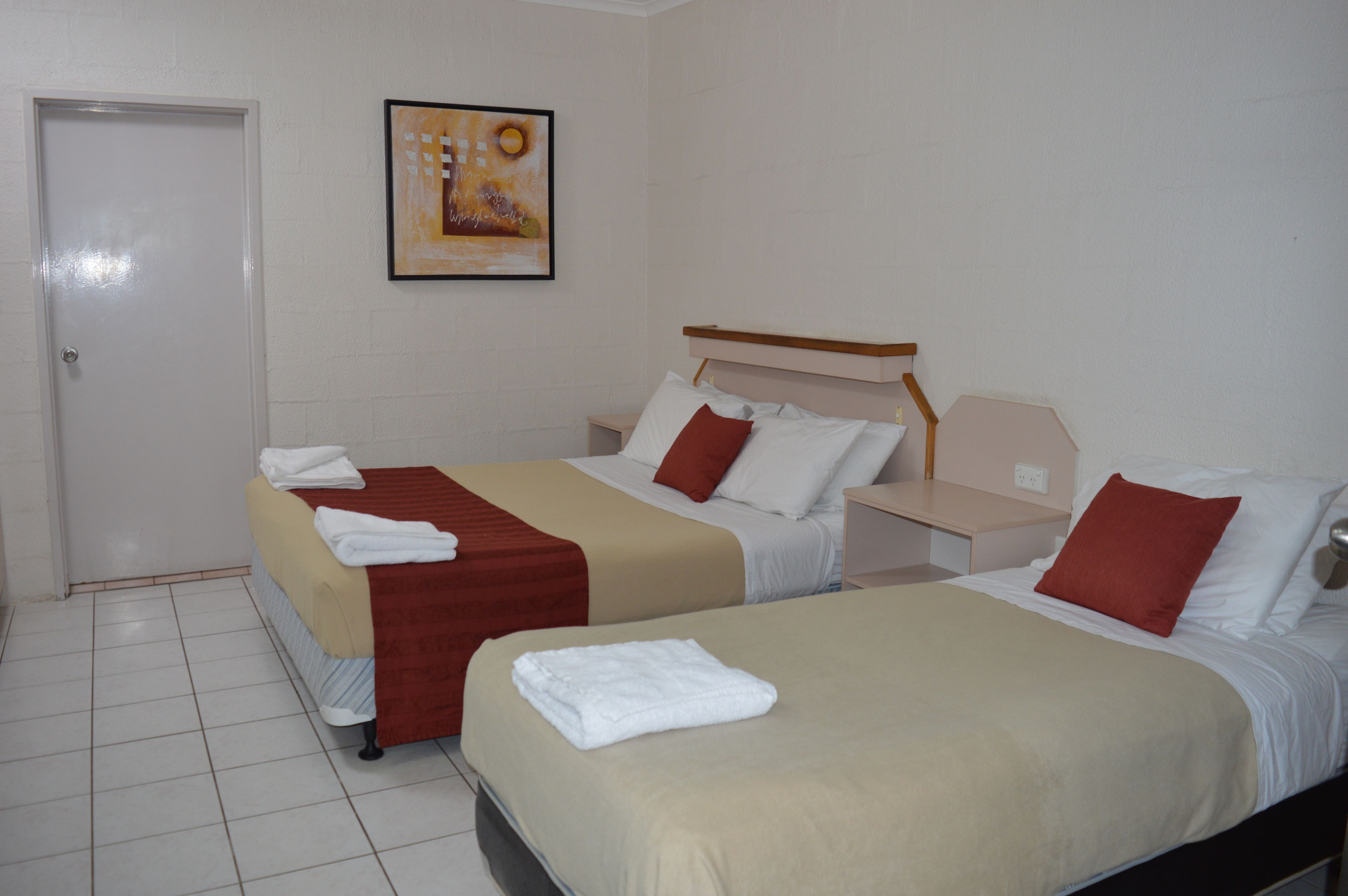 A hotel room with two beds, each with a red and white pillow, a beige blanket, and a white towel. There is a framed artwork on the wall and a door to the left.