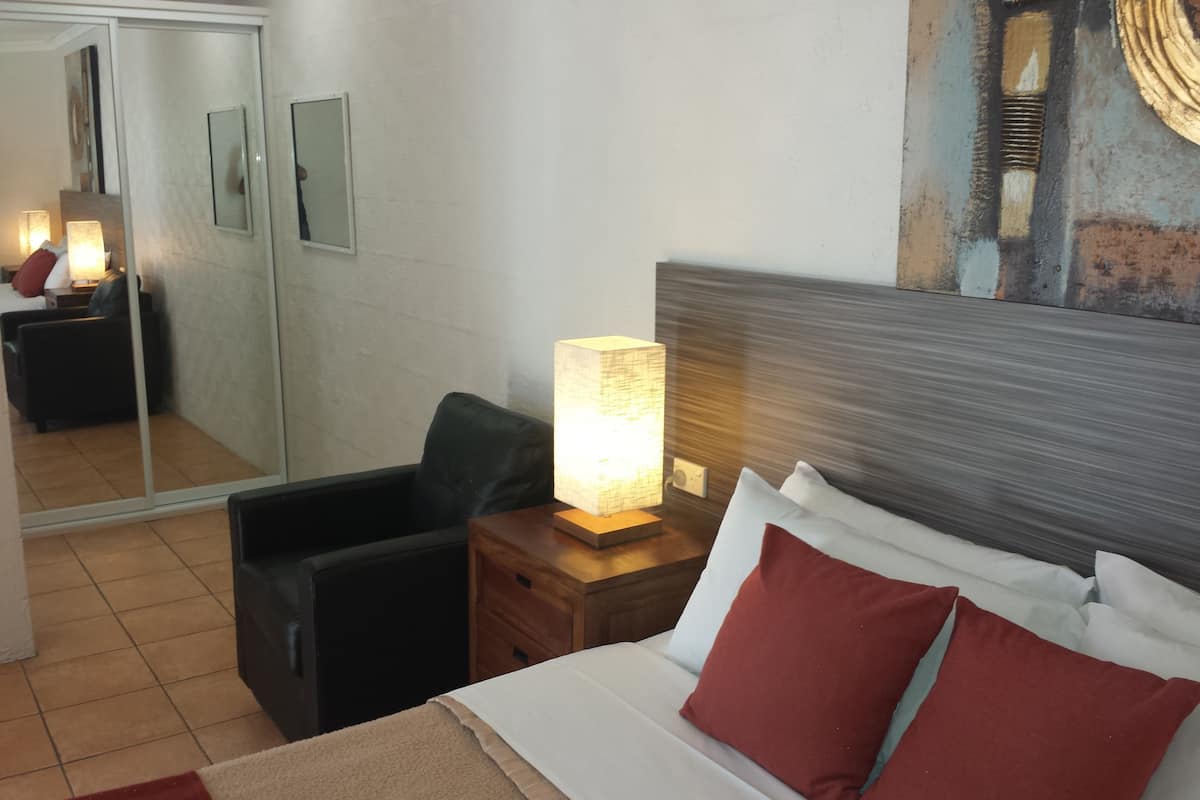 A hotel room with a bed, a nightstand with a lamp, a chair, a mirror, and a wall with a textured design.