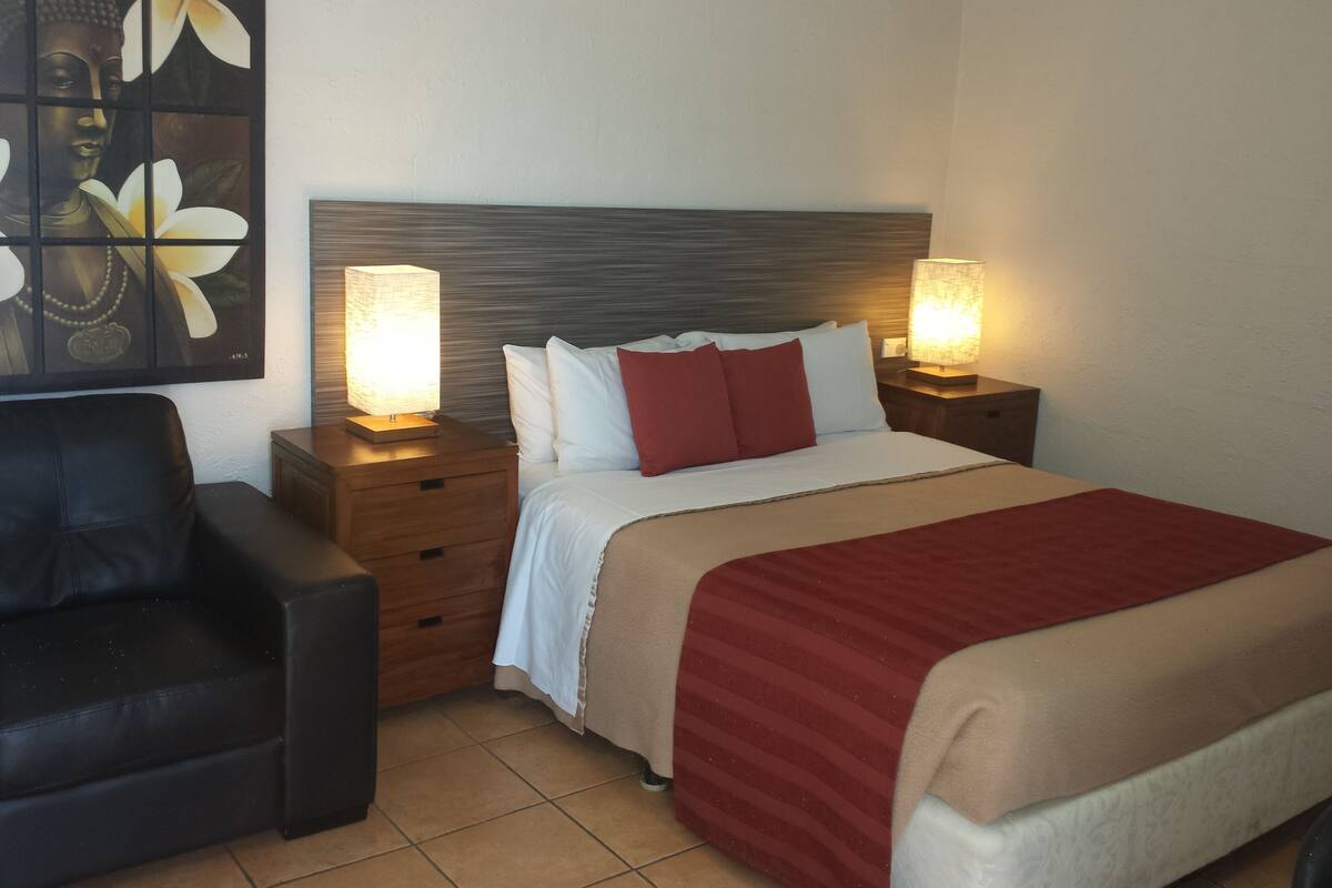 A hotel room with a bed, two bedside lamps, a wooden nightstand, a black leather chair, and a wall-mounted artwork.