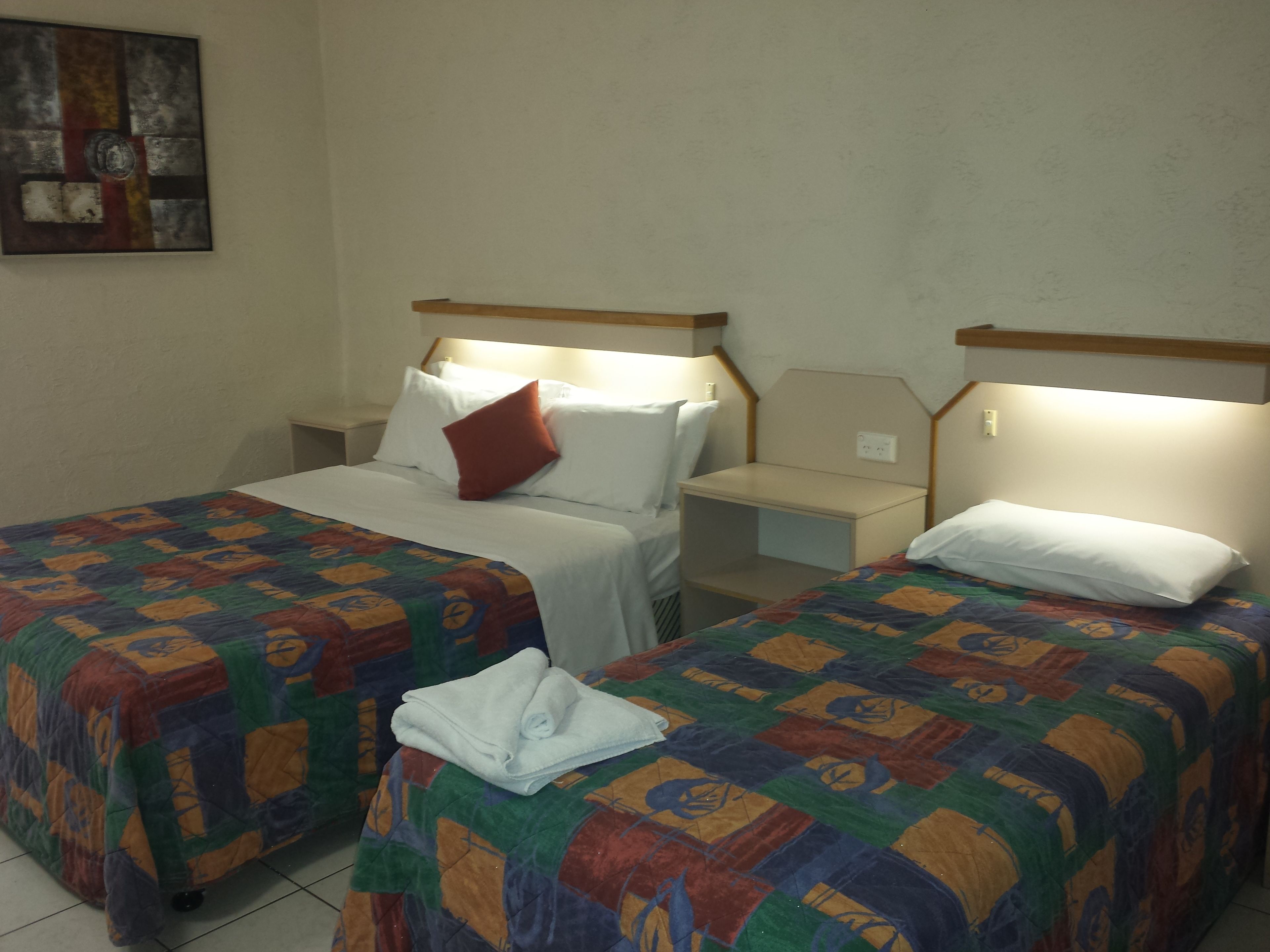 A hotel room with two single beds, each with a colorful quilt, a nightstand, and a bedside table.