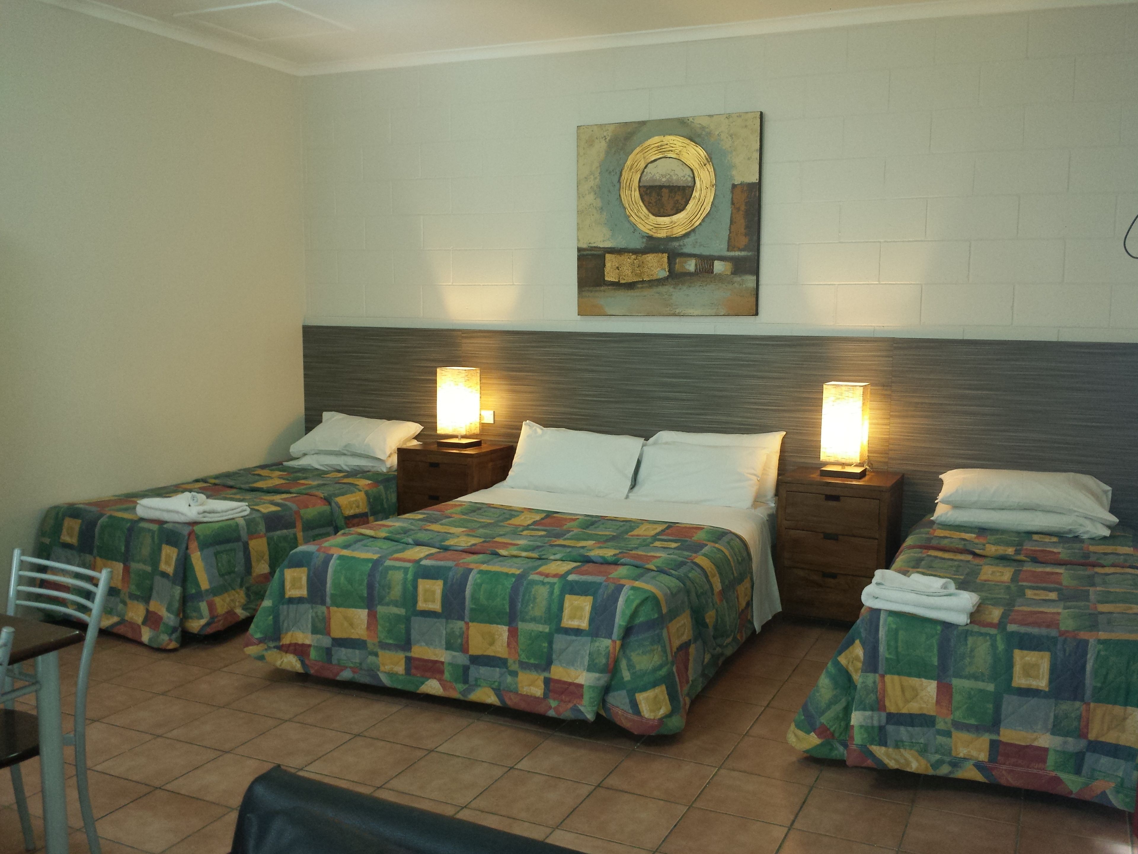 Habitación de hotel con dos camas individuales, una cama doble y una silla. Hay dos mesitas de noche con lámparas, un cuadro en la pared y suelo de baldosas.