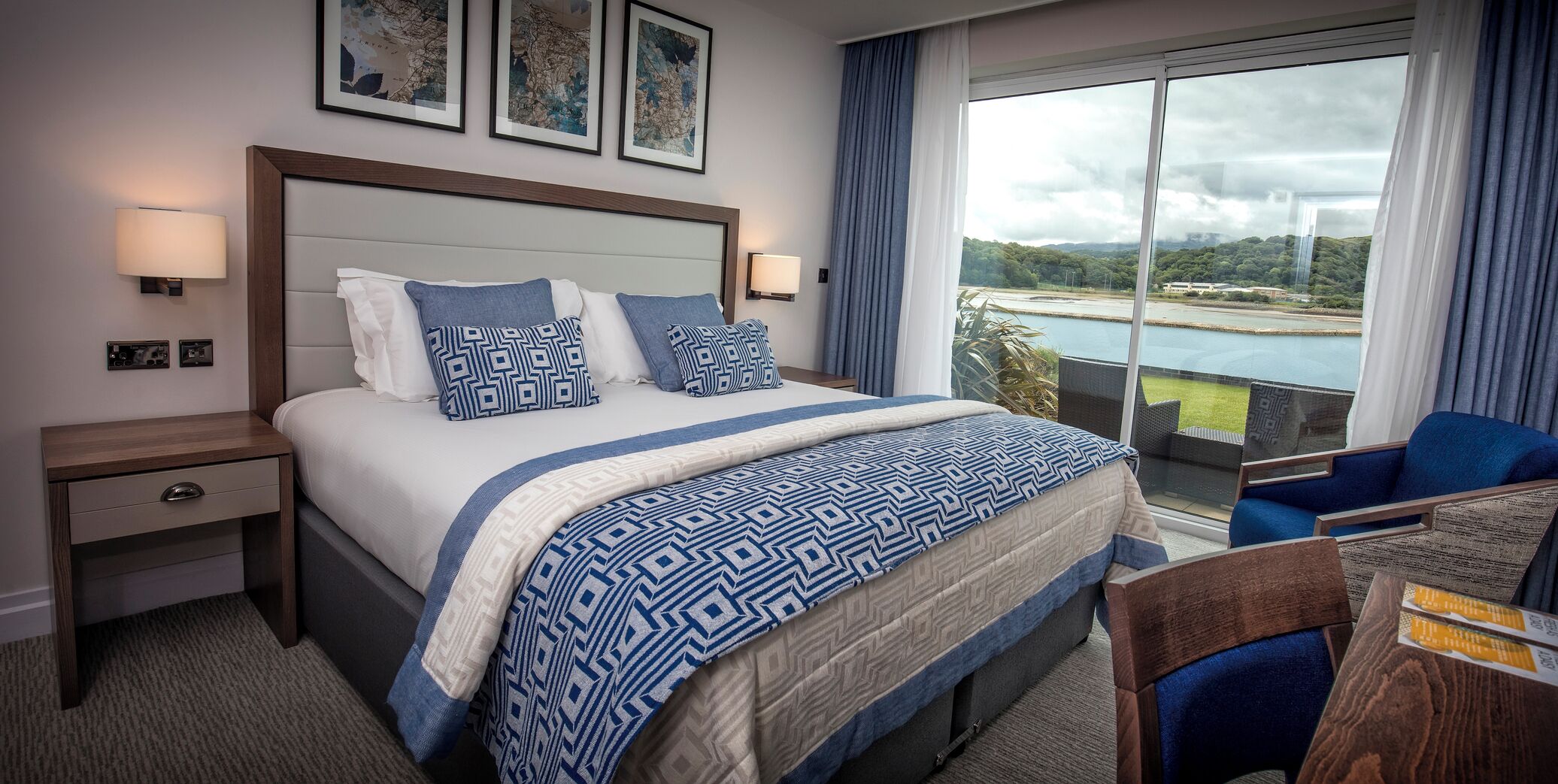 A hotel room with a large bed, a nightstand, a desk with a chair, and a balcony with a view.