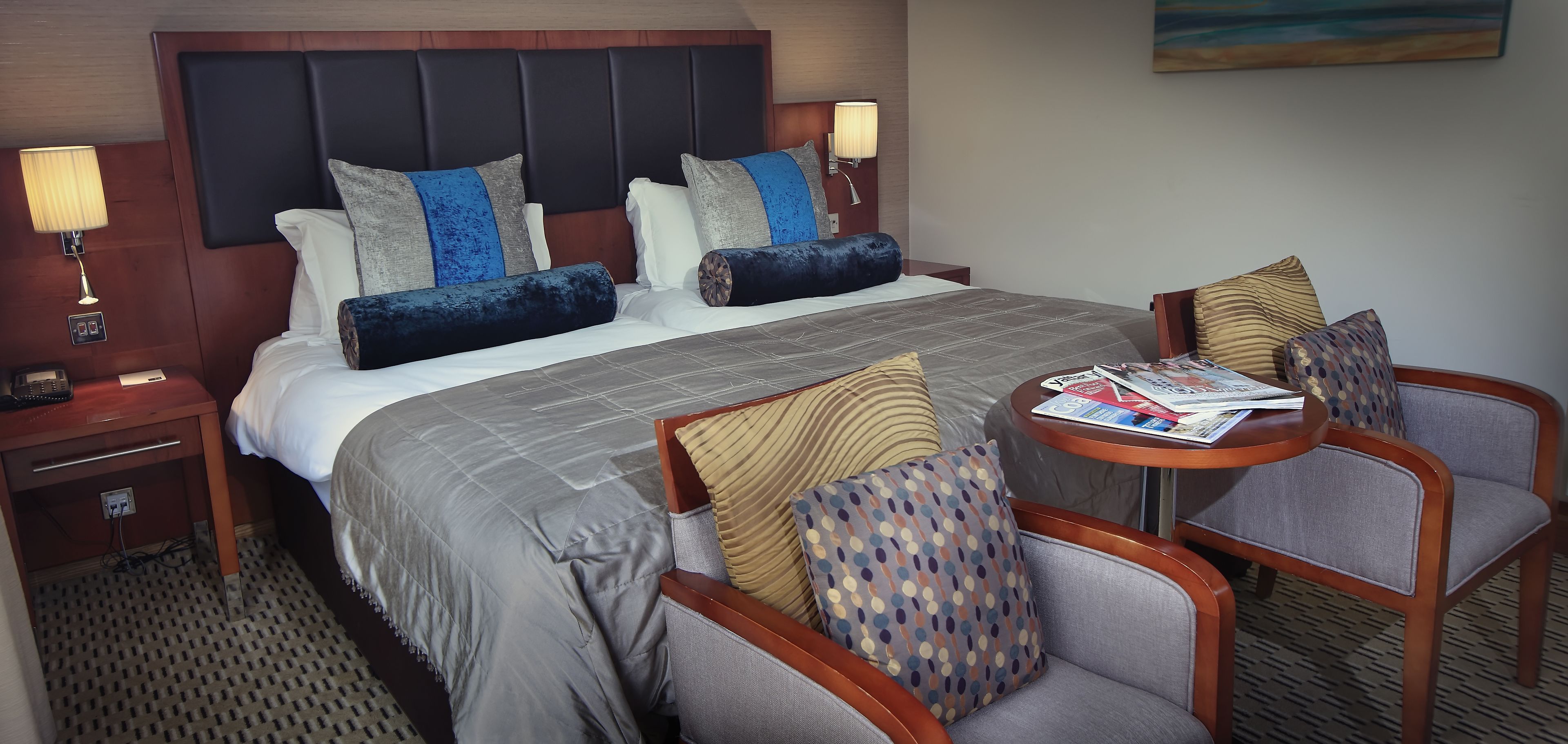 A hotel room with a large bed, two bedside tables, a nightstand, a round table with chairs, and a painting on the wall.
