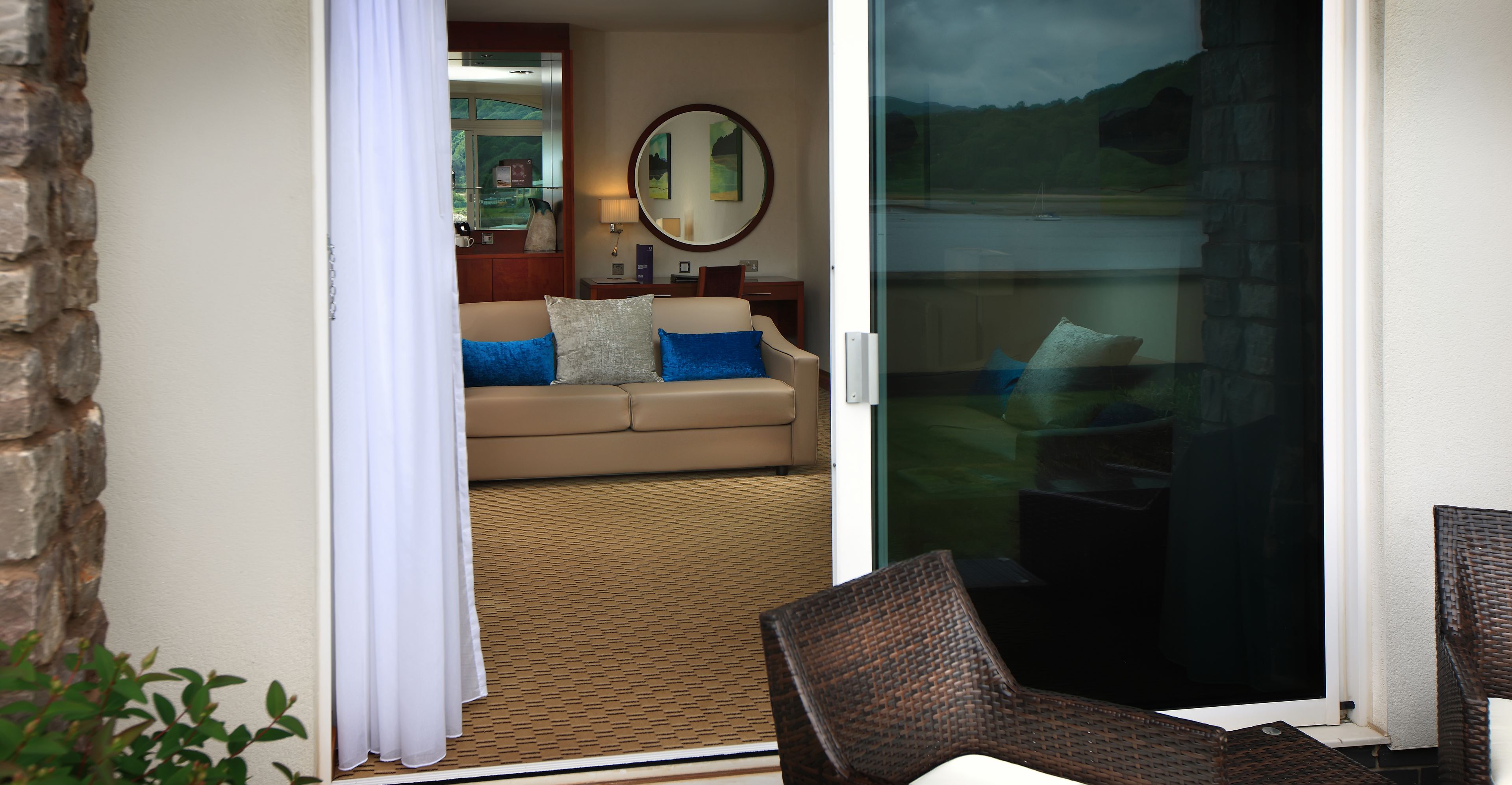 A room with a sliding glass door leading to a balcony, a beige sofa with blue cushions, a round mirror on the wall, and a wicker chair on the balcony.
