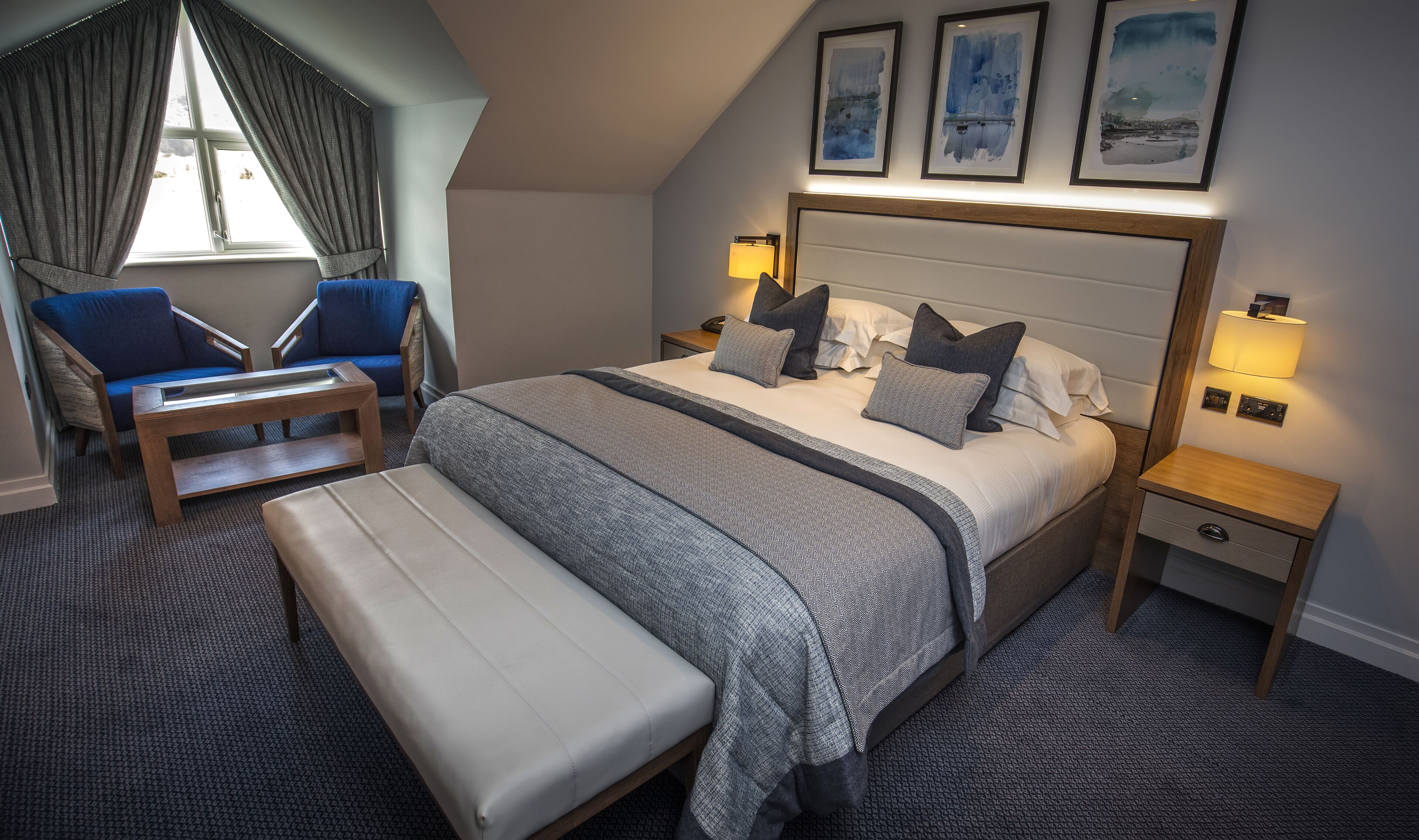 A hotel room with a large bed, a bedside table with a lamp, a chair, a bench, and a window with curtains.