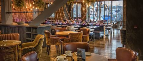 Un restaurante con mesas redondas y sillas acolchadas, una zona de bar con mostrador de madera, y vistas a un puerto deportivo a través de amplias ventanas.