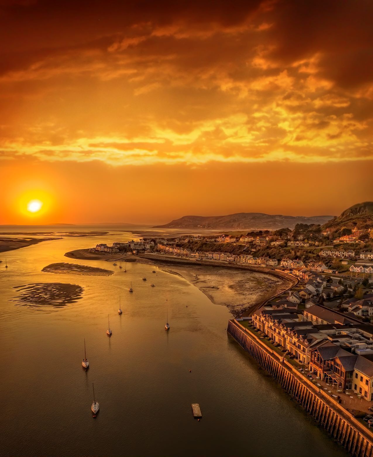 A coastal town at sunset with boats in the water, a pier, and buildings along the shore.