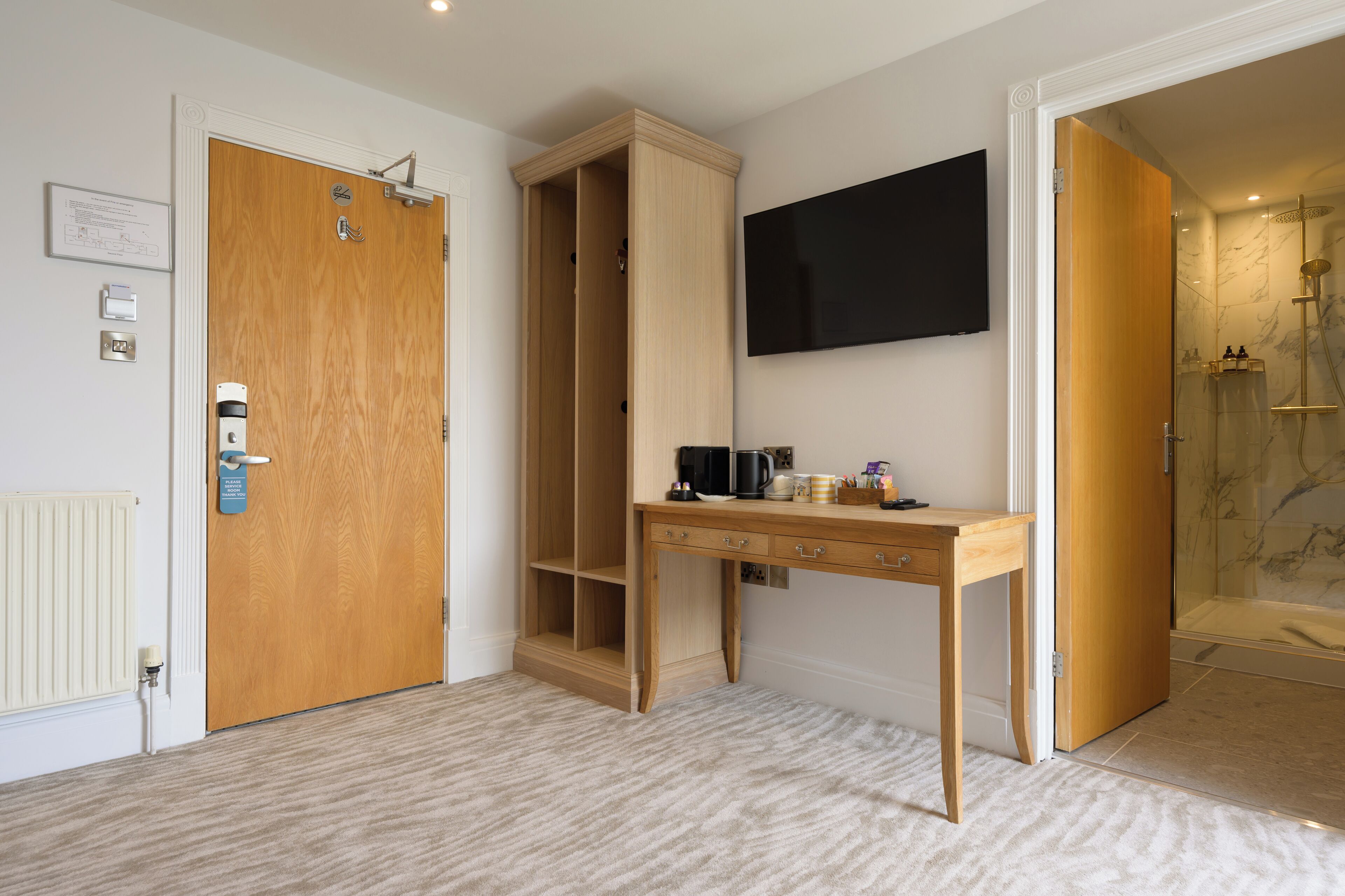 A hotel room with a wooden door, a desk with drawers, a mounted television, and a shower area.