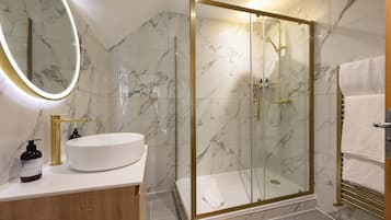 A modern bathroom with a glass-enclosed shower, a marble-tiled wall, a round mirror, and a wooden vanity.