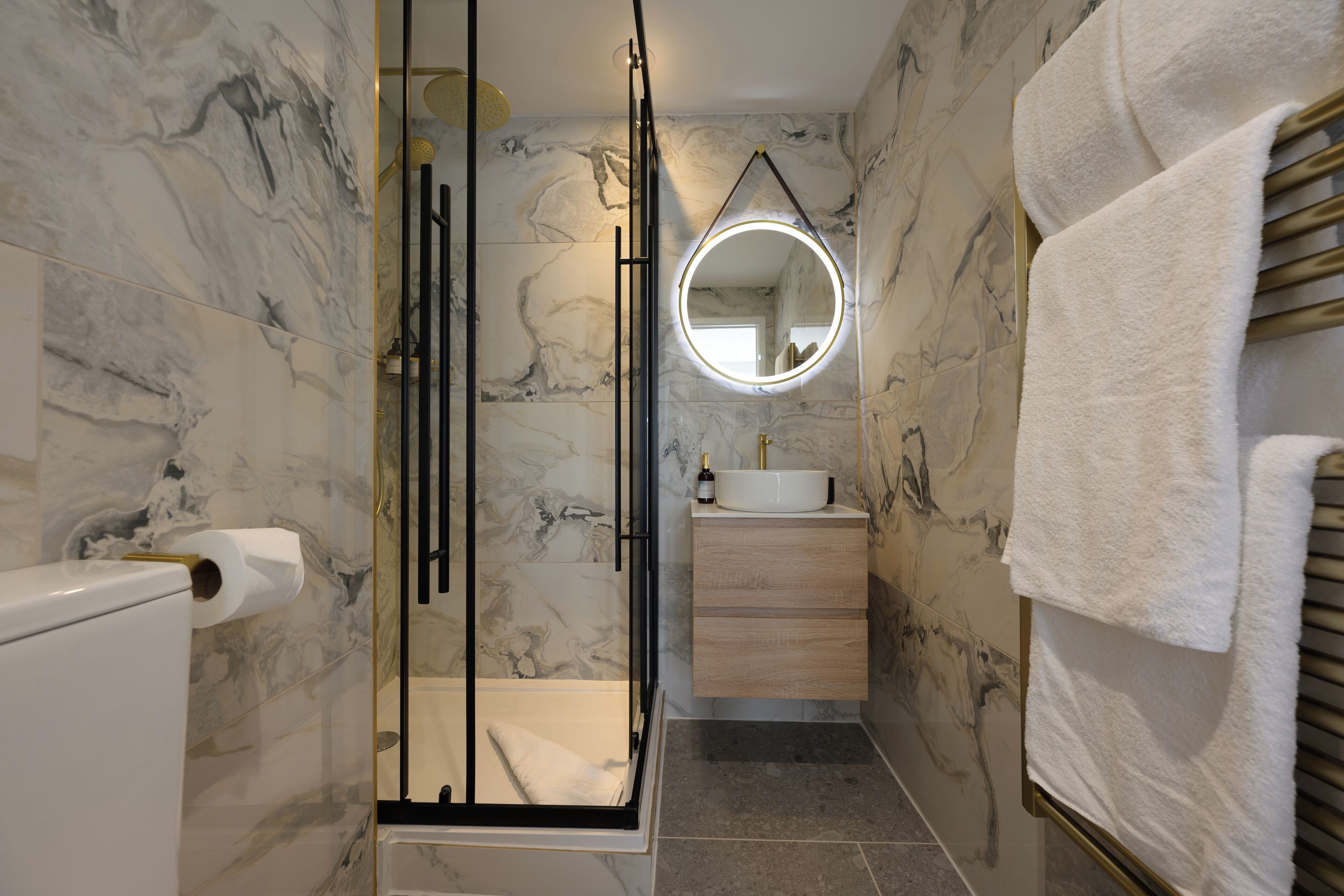 A bathroom with marble walls, a glass shower enclosure, a round mirror, a sink, and a towel rack.