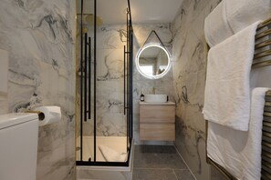 A bathroom with marble walls, a glass shower enclosure, a round mirror, a sink, and a towel rack. - The Old Quay House Hotel (Fowey)
