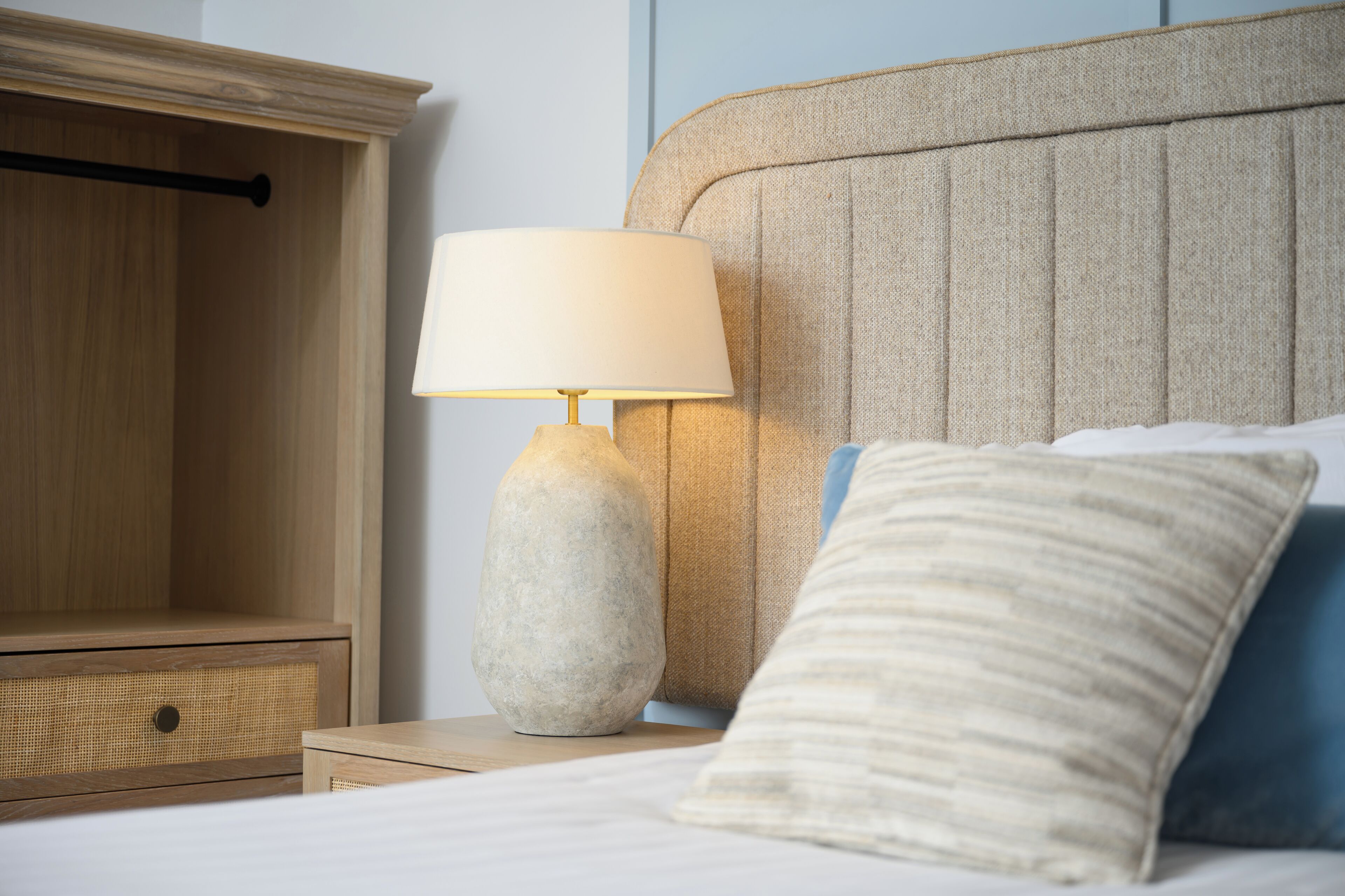 Una cama bien hecha con cabecera, una lámpara de noche y una mesita de noche de madera con cajón.