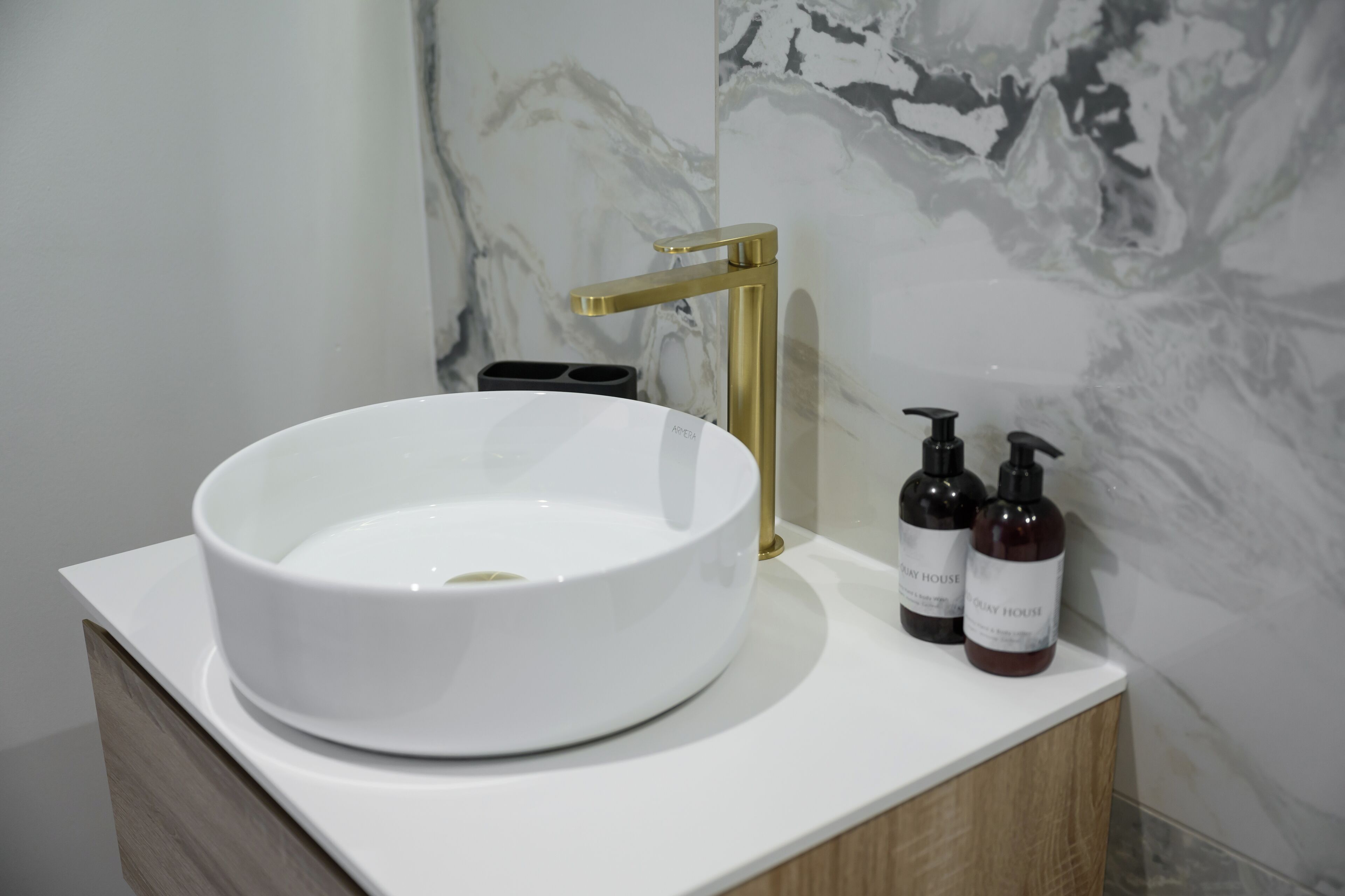 A bathroom with a white sink, gold faucet, and two bottles of shampoo on a white countertop.