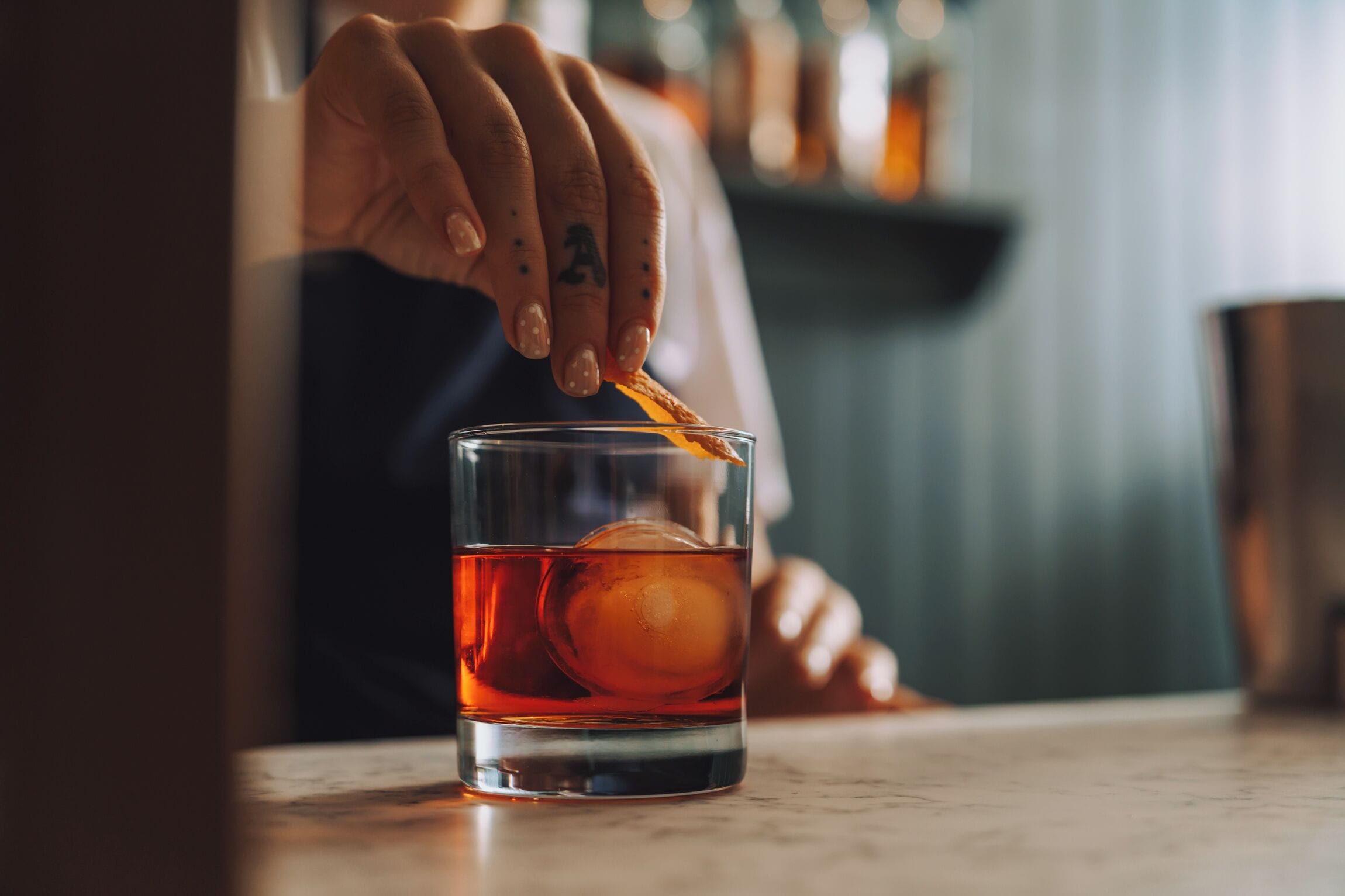En hånd placerer et appelsinstykke i et glas med en gylden væske, sandsynligvis en cocktail, med en uskarpt baggrund, der antyder en bar eller restaurant.