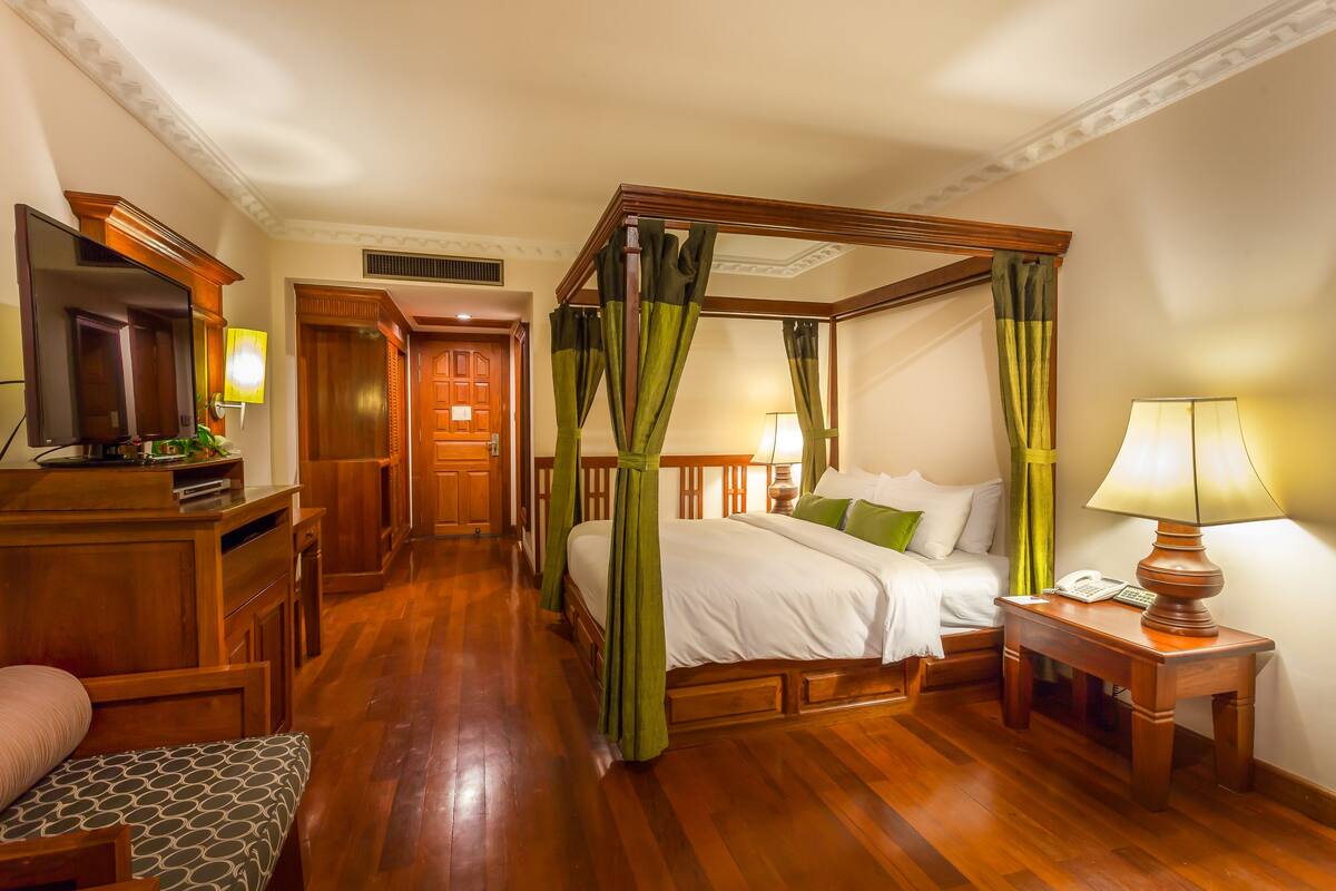 Un letto a baldacchino con tende verdi, un comodino in legno con una lampada, un tavolino da notte con un telefono e una televisione su un mobile in legno.