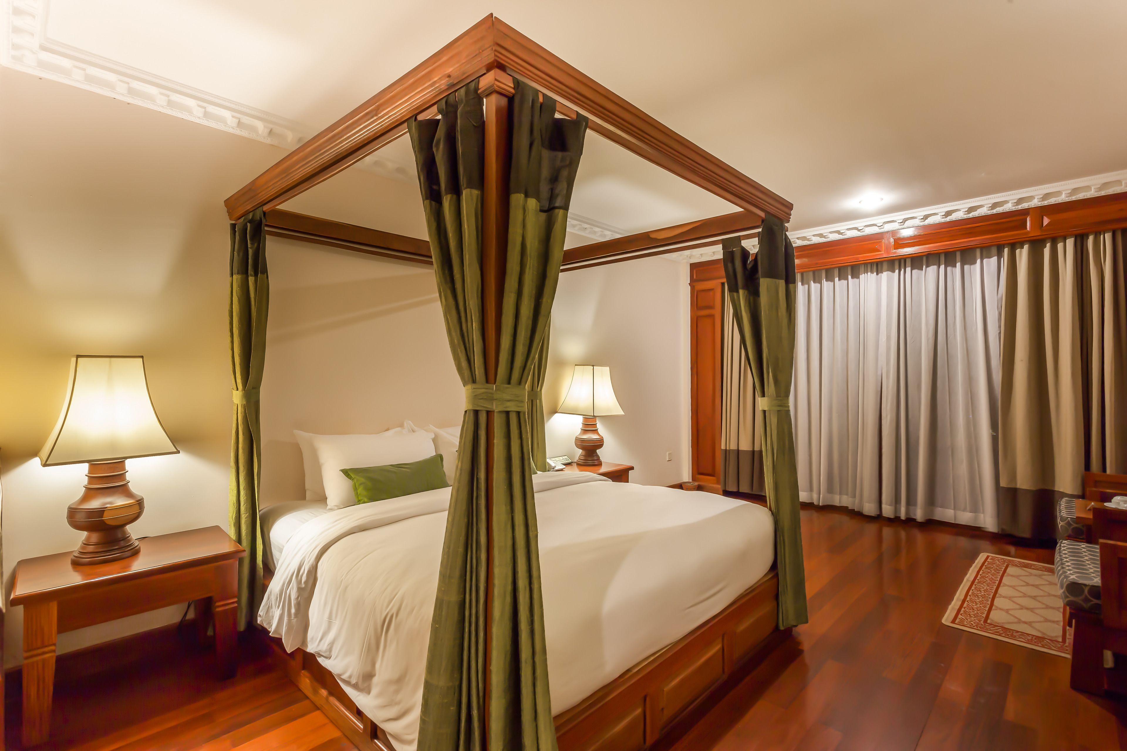 A four-poster canopy bed with a wooden frame, two bedside tables with lamps, a nightstand, and a wooden floor.