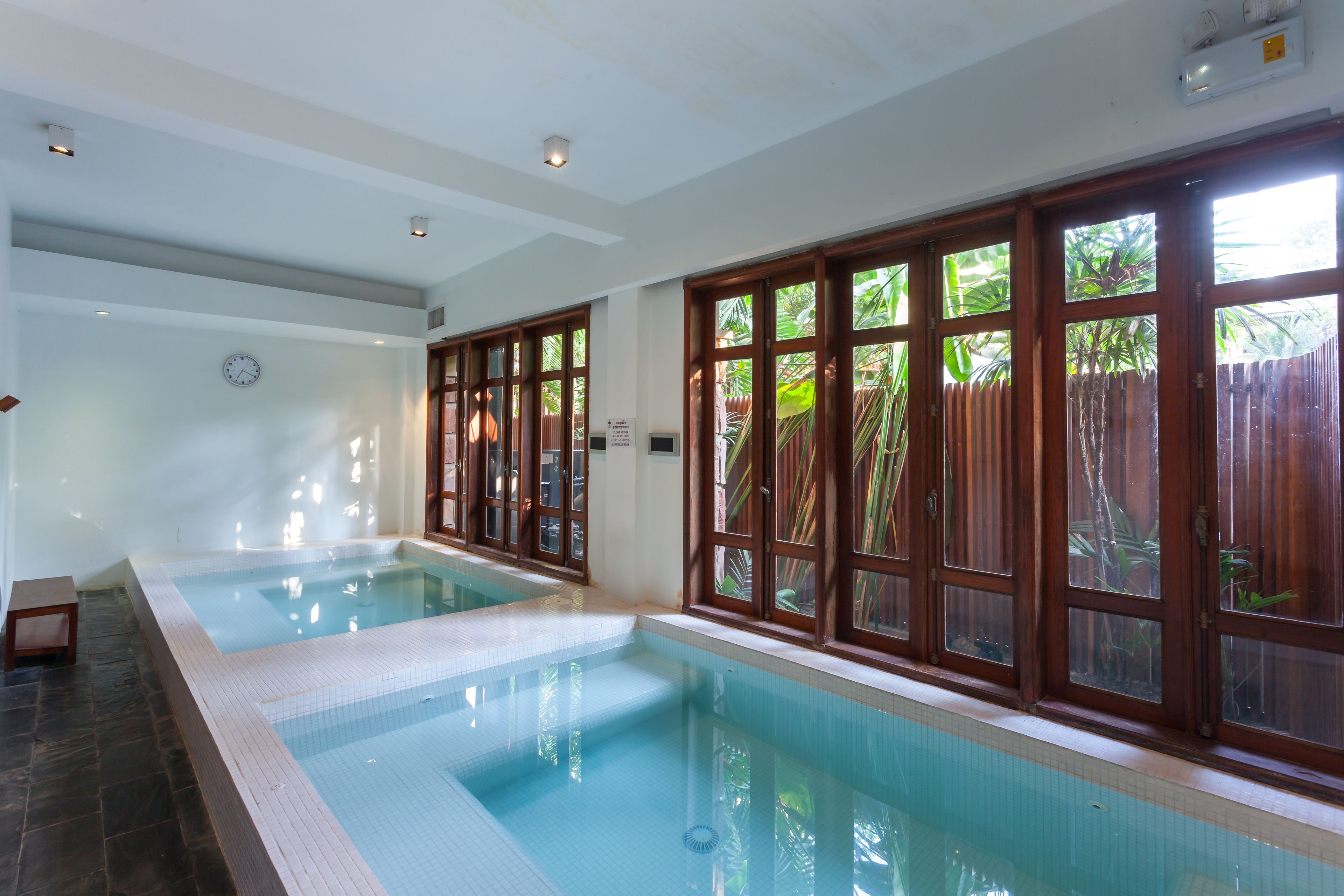 An indoor swimming pool with clear glass walls, a tiled floor, and a wooden fence visible through the windows.