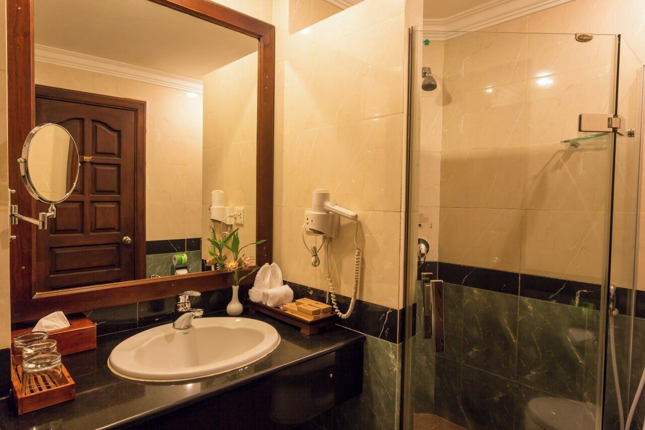 Une salle de bain d’hôtel avec un grand miroir, un lavabo, un porte-serviettes, une cabine de douche en verre et une porte avec une poignée.