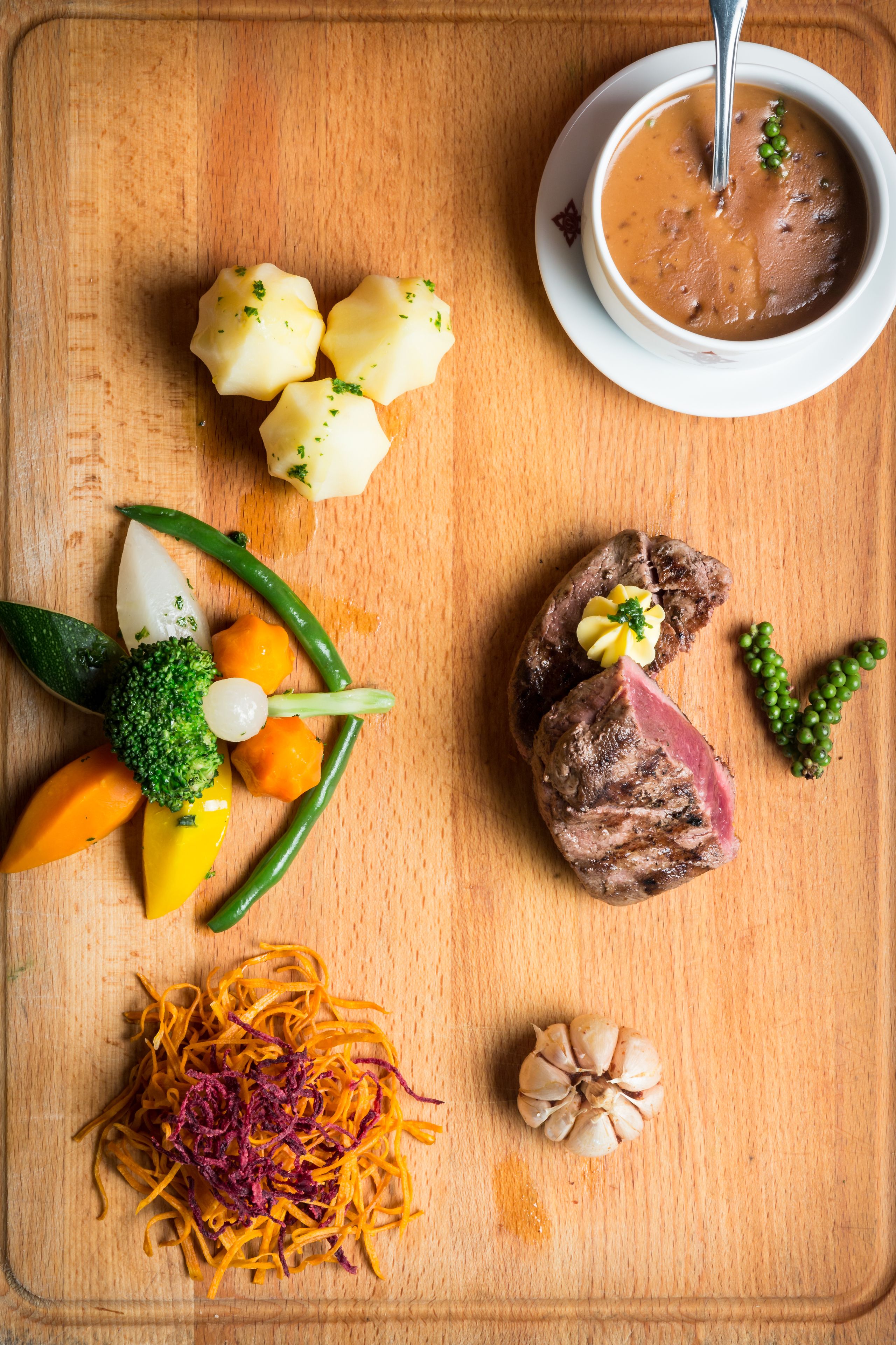 A wooden cutting board with a variety of food items including potatoes, broccoli, carrots, a piece of meat, and a bowl of soup.