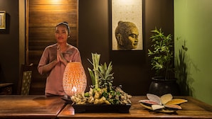 Una persona de pie frente a un mostrador de recepción de madera con una bandeja de jengibre y otras especias, una lámpara decorativa y una estatua de Buda en la pared.