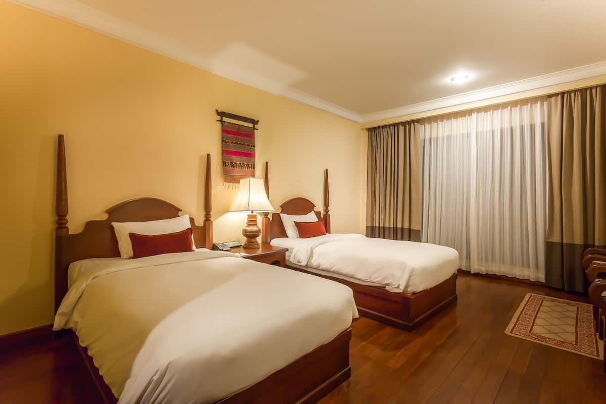 Camera d'albergo con due letti in legno, un comodino con lampada, un quadro appeso al muro e un'ampia finestra con tende.