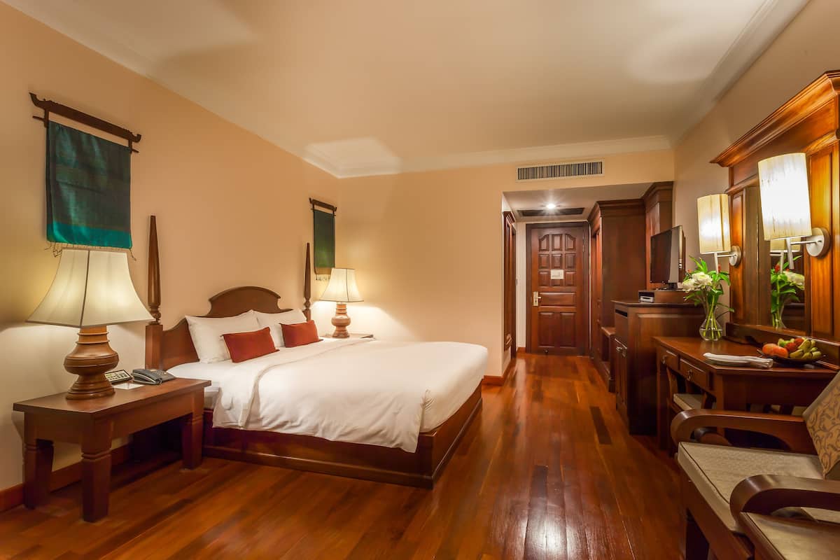 Camera d'albergo con un letto in legno, due lampade sui comodini, un comodino con un telefono, una scrivania in legno con un vaso di fiori e uno specchio.