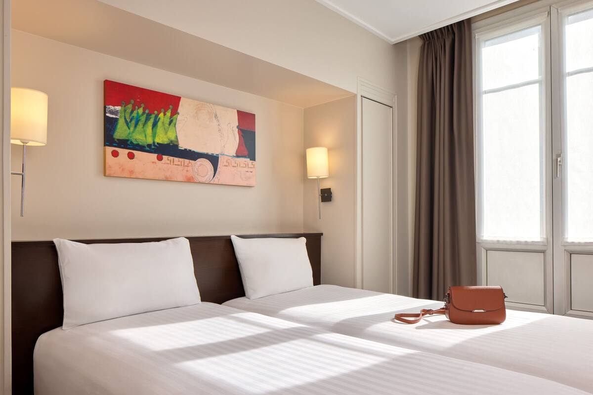 Una camera d'albergo con un letto grande, due abat-jour, una finestra con tende e un quadro astratto dai colori vivaci appeso alla parete.