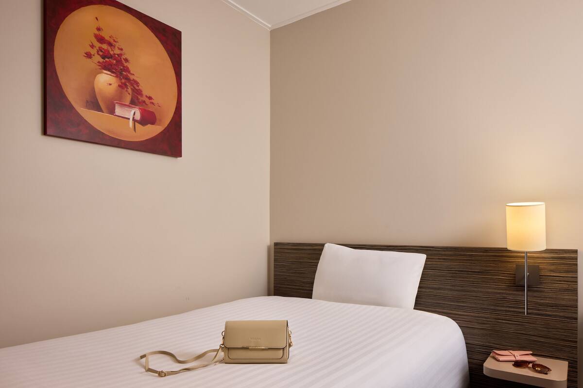 Una camera d'albergo con un letto, un comodino con una lampada, un'opera d'arte appesa al muro e un tavolino con una borsa e un oggetto rosa.