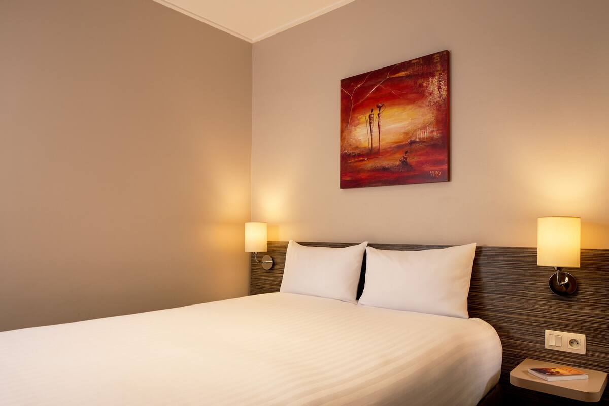Una camera d'albergo con un letto grande, due abat-jour, un quadro appeso al muro e un comodino con un libro.