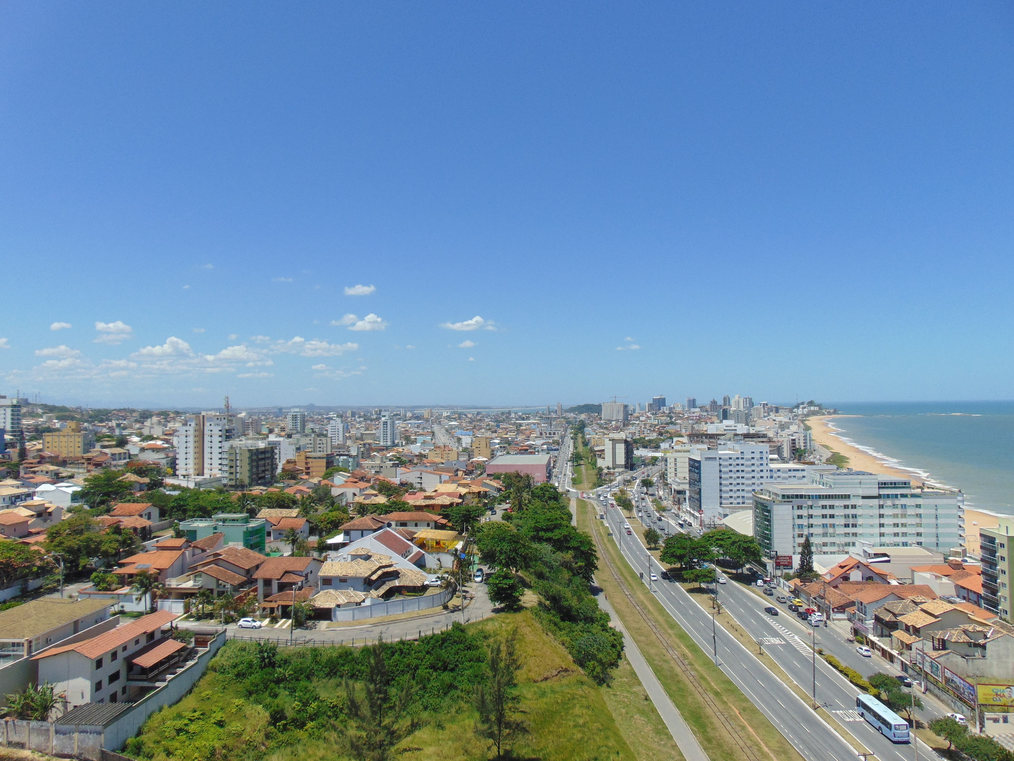 Un paysage urbain côtier, avec un mélange de bâtiments résidentiels et commerciaux, une large route et une plage au loin.