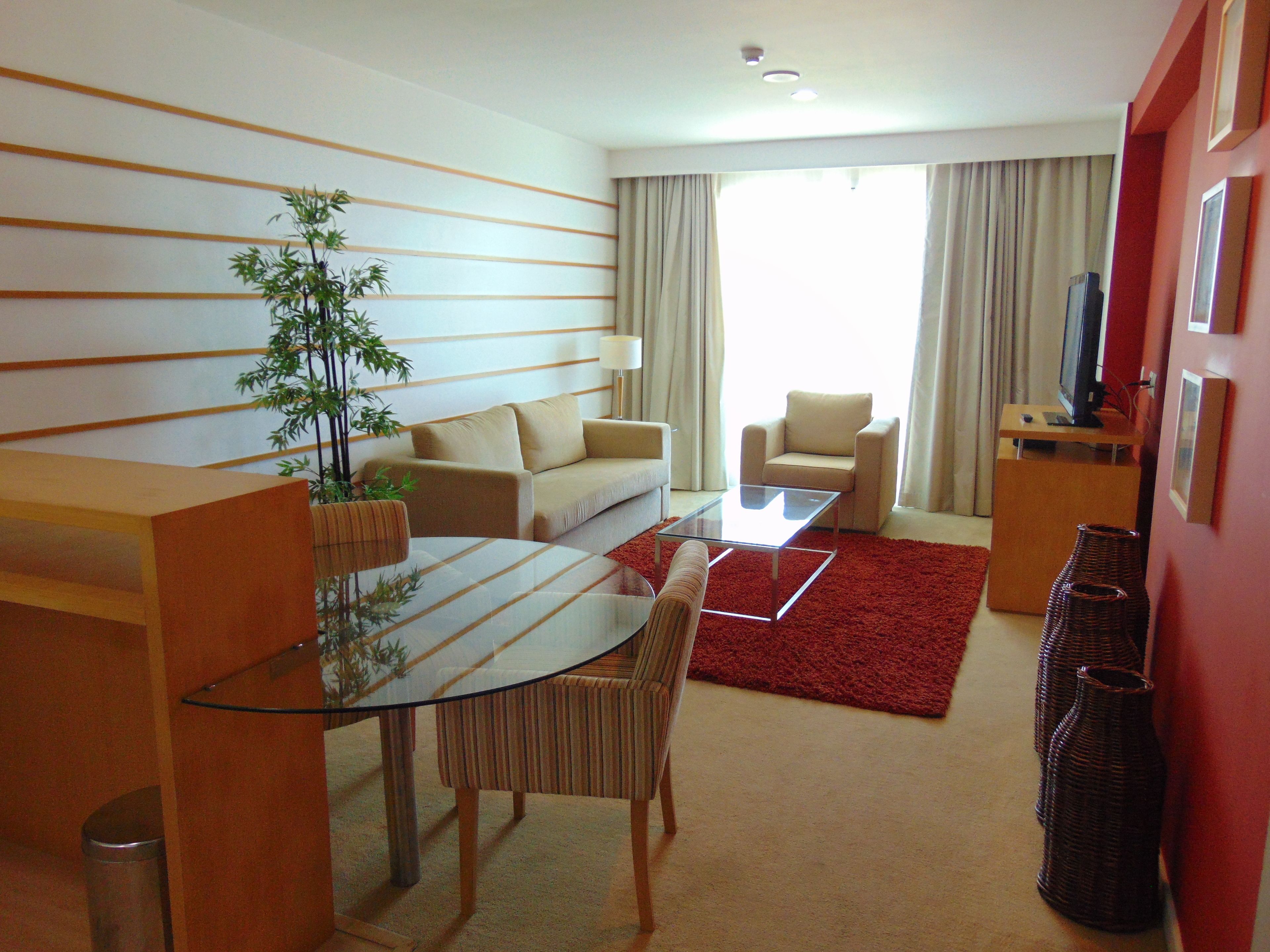 Una sala de estar moderna con un sofá, sillones, una mesa de centro de vidrio, una mesa de comedor con sillas, un tapete rojo y un televisor sobre un mueble.
