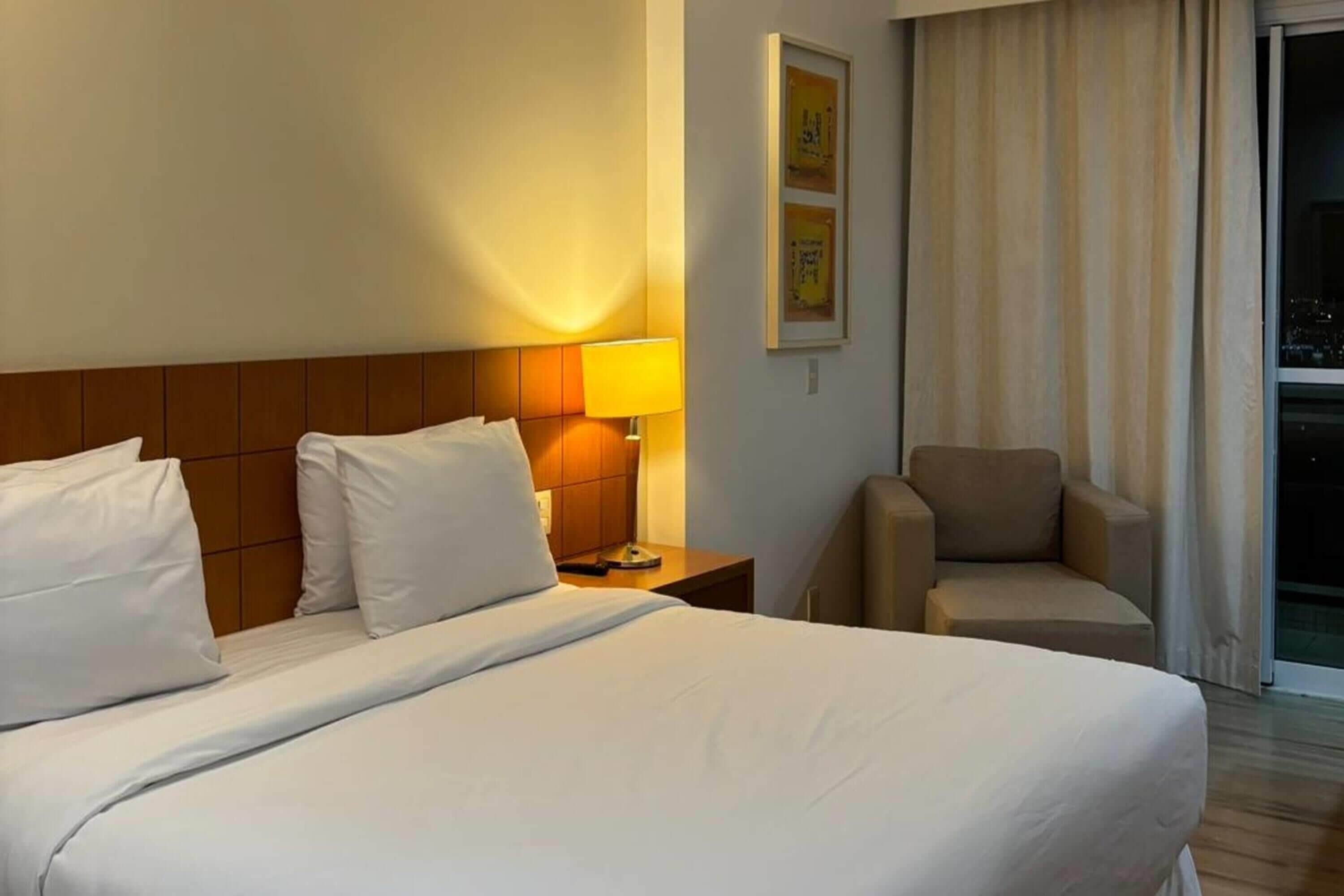 A hotel room with a large bed, a nightstand with a lamp, a chair, and a window with curtains.