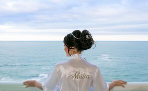 Een persoon in een wit shirt met de tekst 'Noiva' erop geborduurd, staat bij een reling met uitzicht op zee.